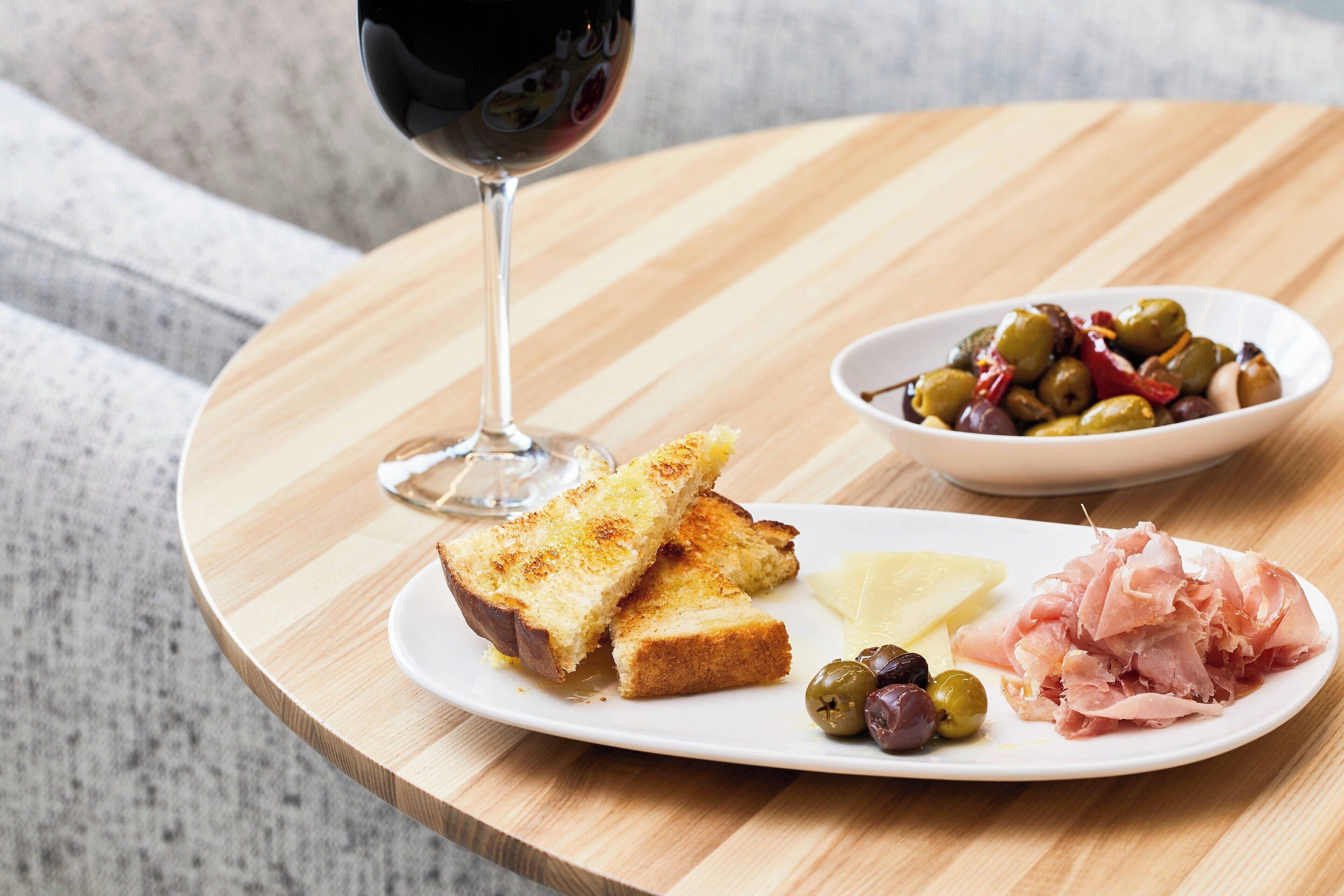 Una mesa de madera con una tostada, aceitunas y jamón, además de una copa de vino tinto.