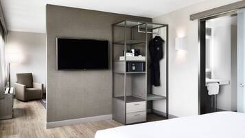 Une chambre d’hôtel moderne dotée d’un grand lit, d’un téléviseur à écran plat fixé au mur, d’une armoire intégrée avec un miroir et d’un petit coin salon avec un fauteuil.