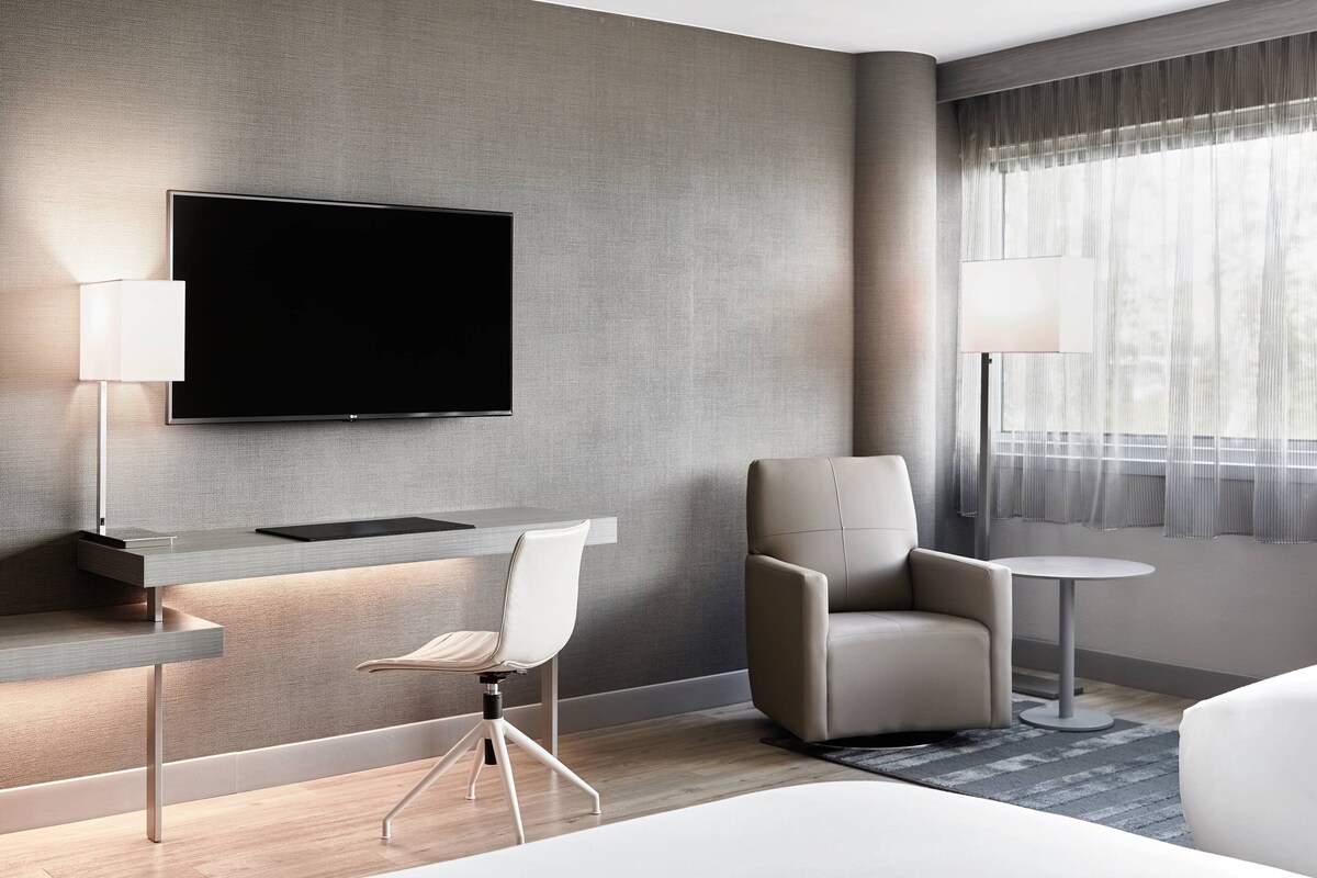 Une chambre d’hôtel moderne équipée d’un téléviseur à écran plat fixé au mur, d’un bureau avec une lampe, d’un fauteuil confortable, d’un canapé et d’une petite table ronde.