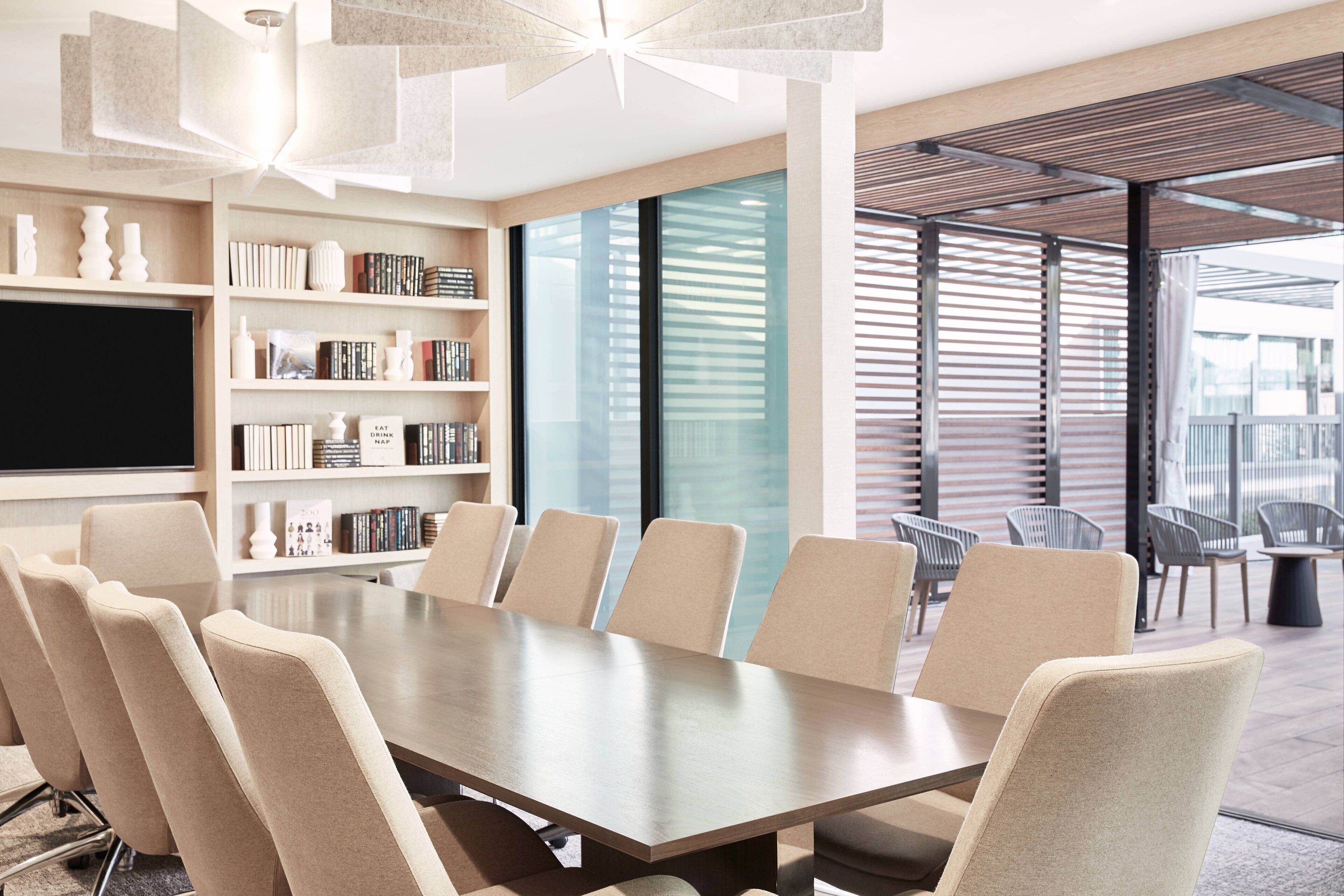 A modern meeting room with a long table, comfortable chairs, a bookshelf, and a large window with blinds.