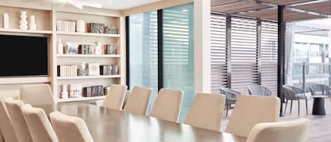 A modern meeting room with a long table, comfortable chairs, a bookshelf, and a large window with blinds.