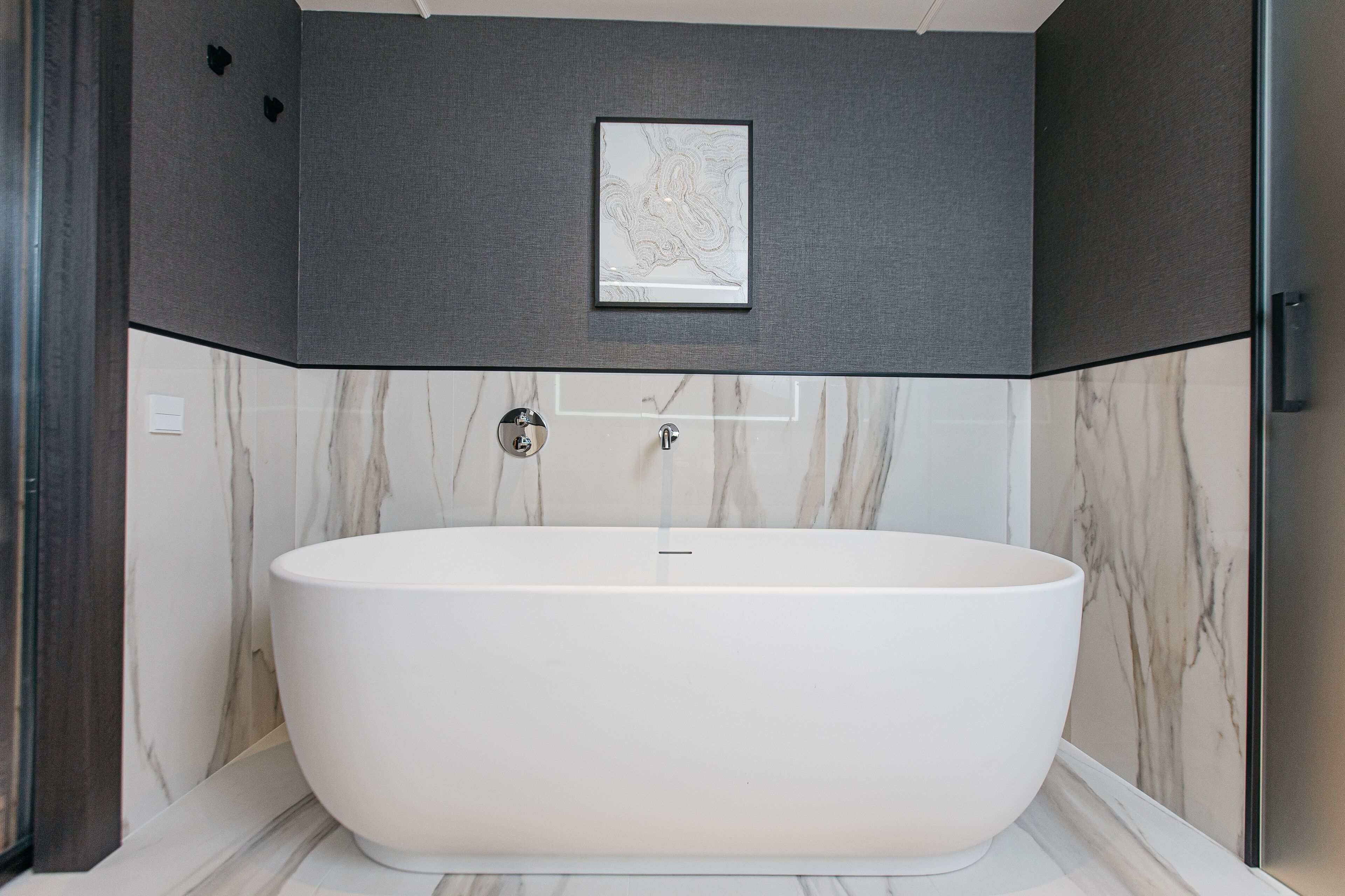 A modern bathroom with a freestanding bathtub, marble walls, and a framed artwork above the tub.