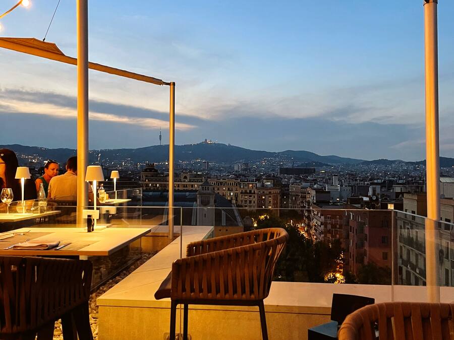 A rooftop terrace with tables and chairs, overlooking a cityscape and mountains.