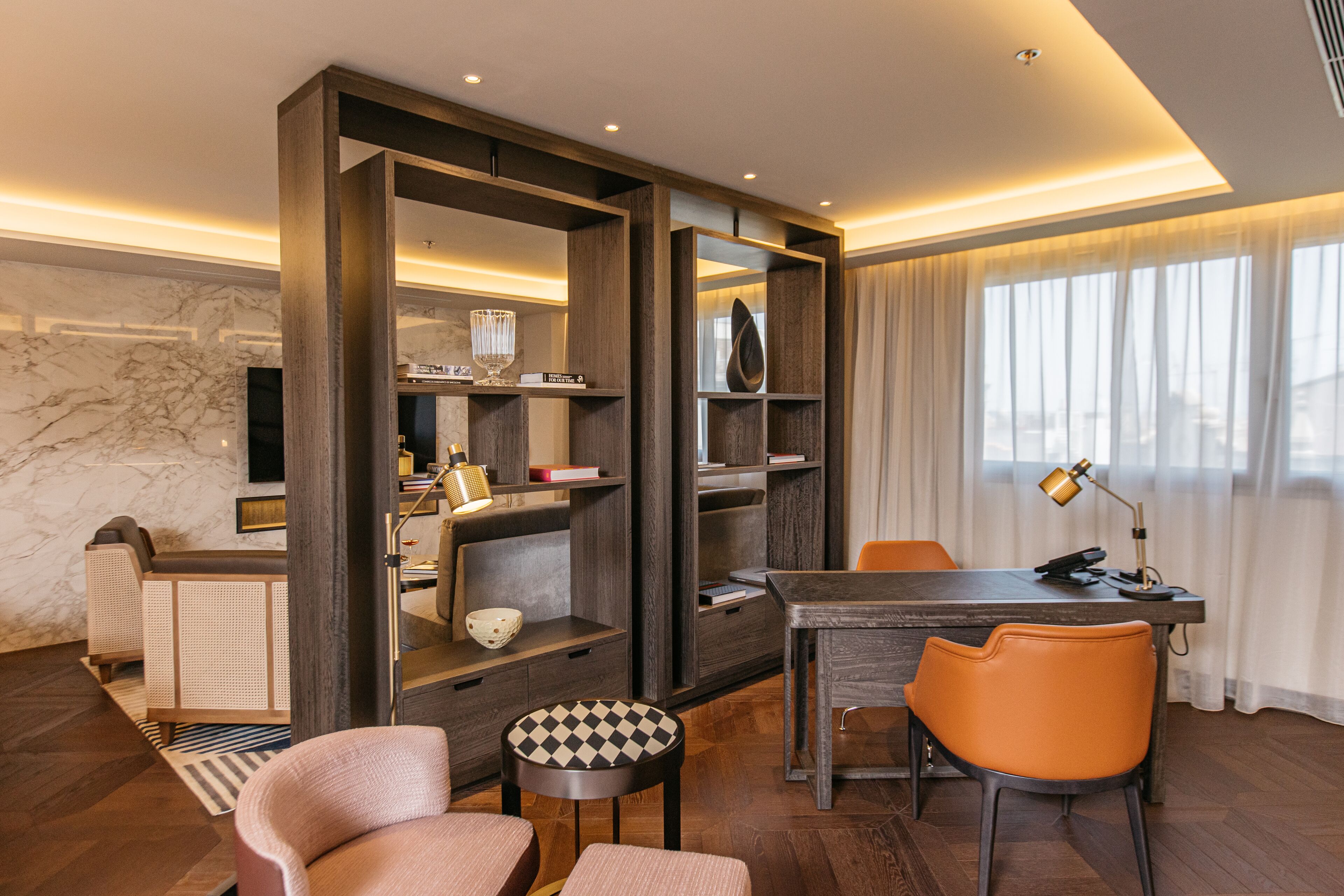 Quarto de hotel moderno com uma secretária de madeira, cadeiras laranja, uma mesa redonda e uma estante.