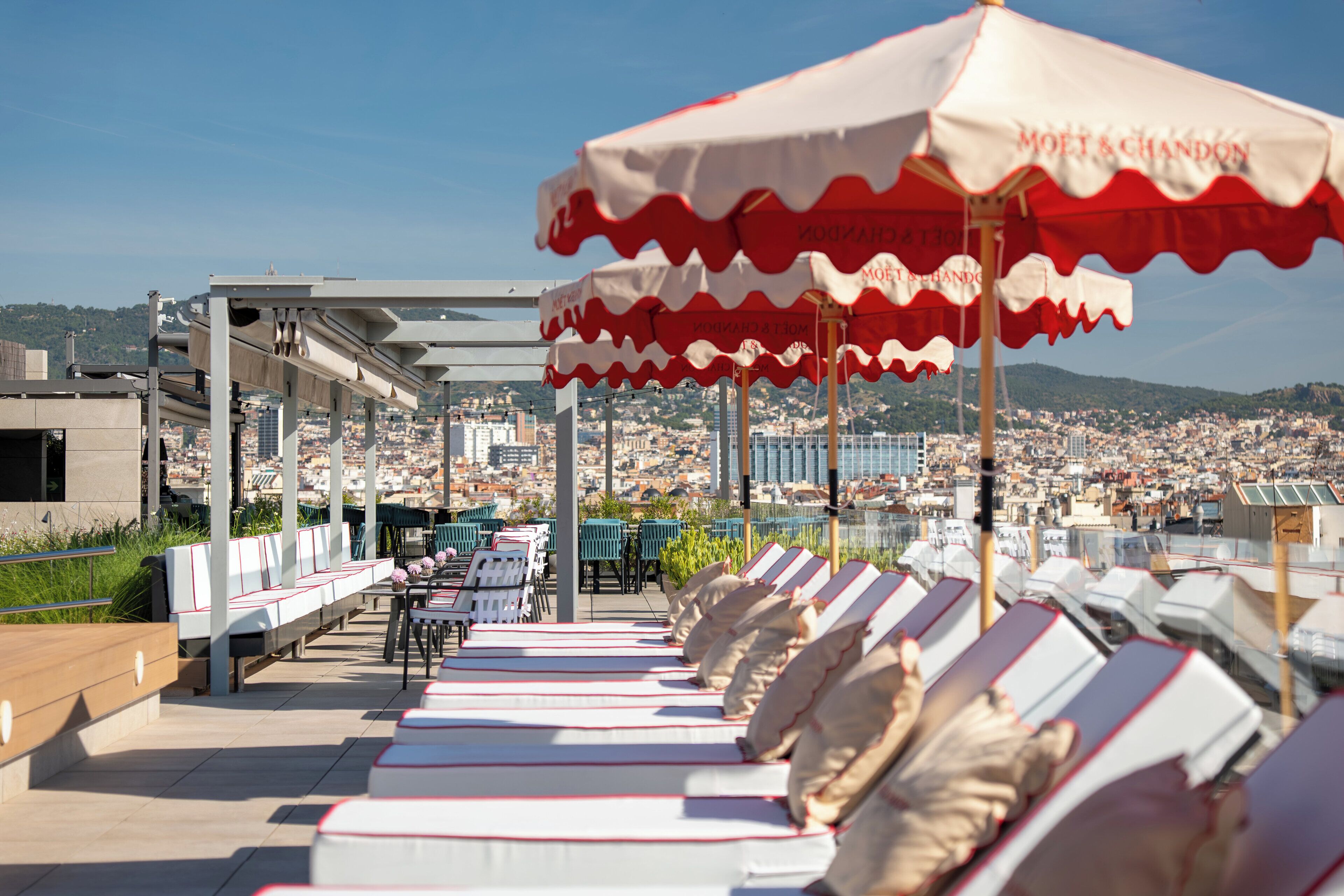 A rooftop terrace with sun loungers and umbrellas, overlooking a cityscape.