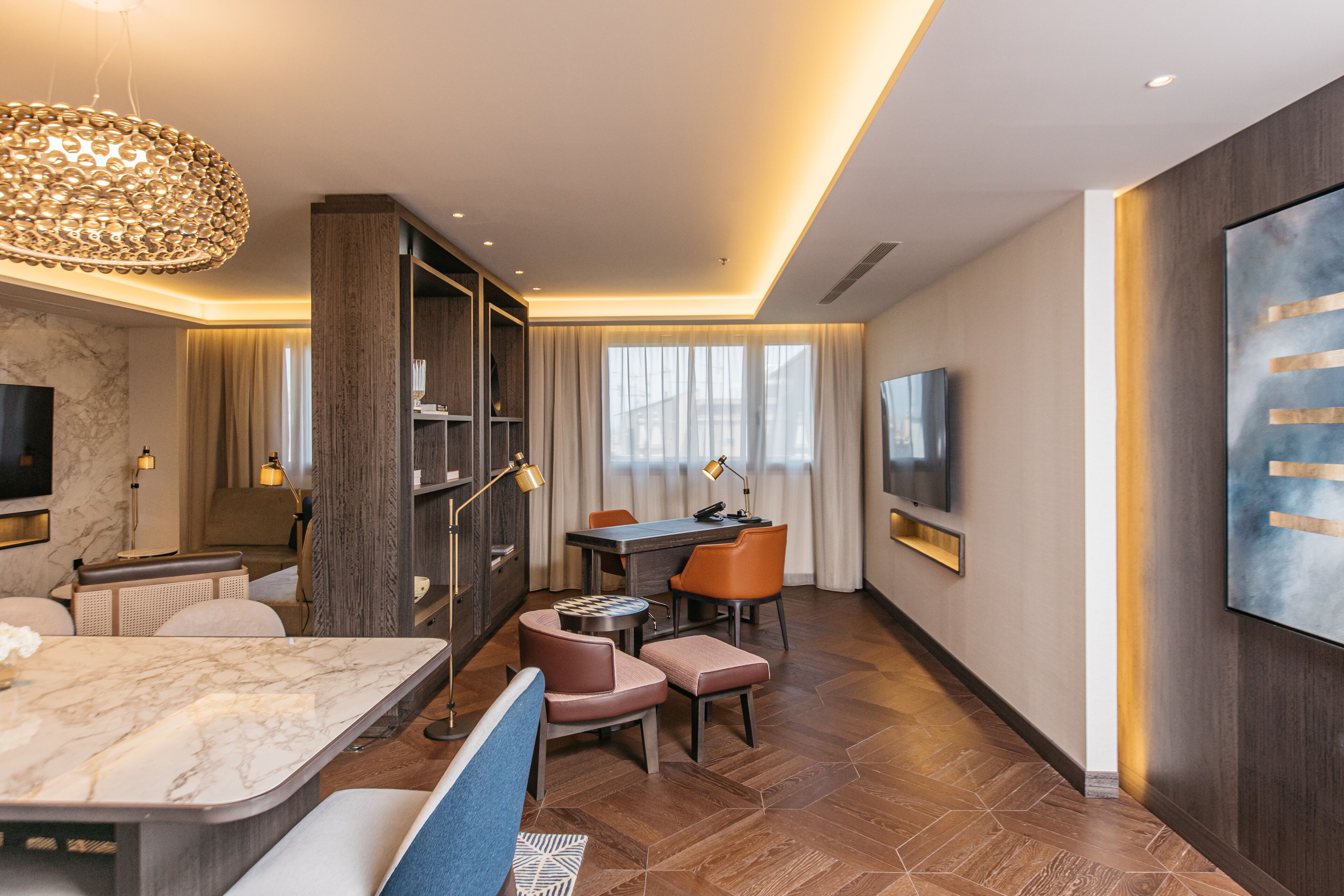 A modern hotel room with a marble-topped dining table, a sofa, a TV mounted on the wall, and a large abstract painting.