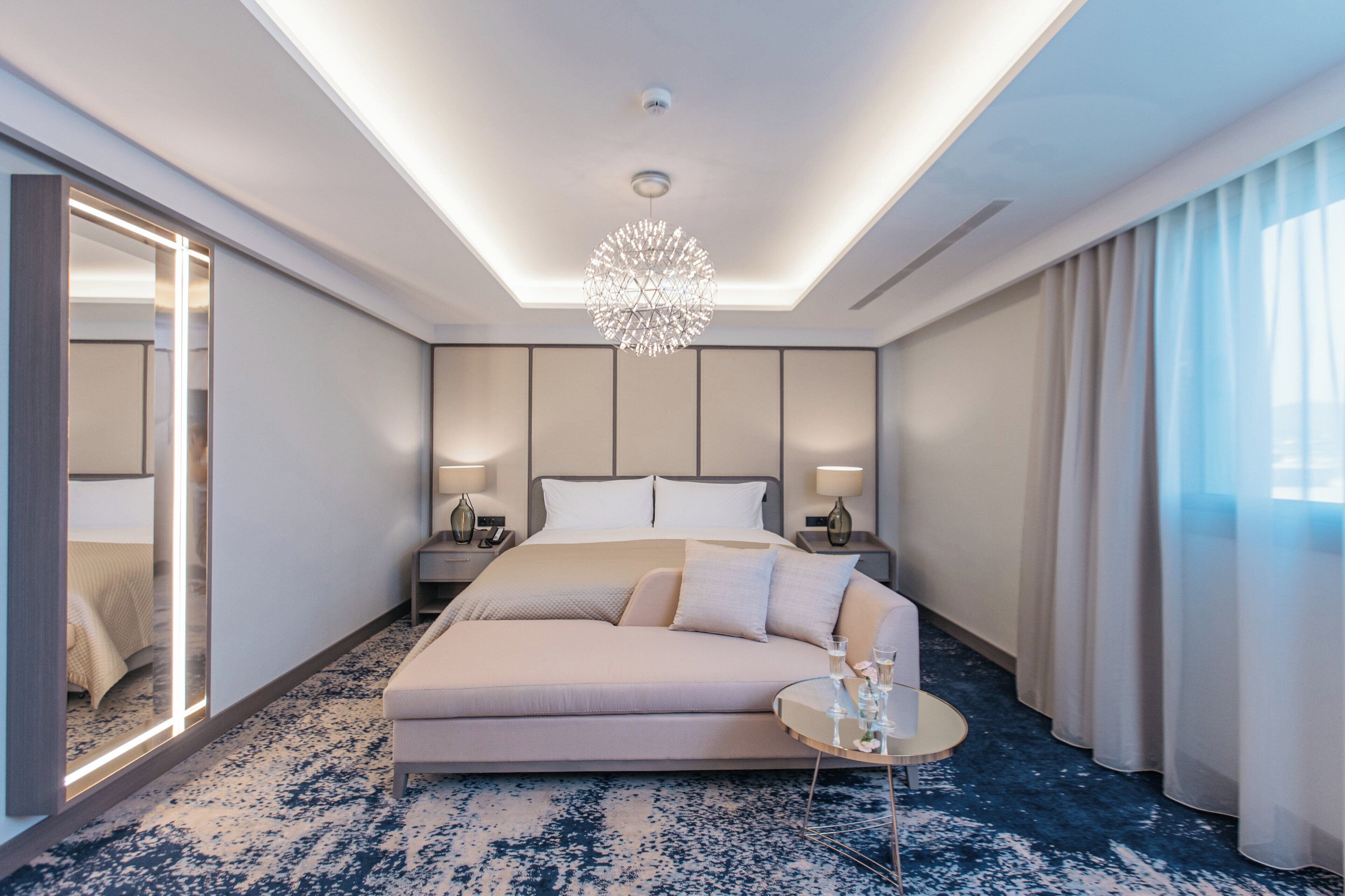A modern hotel room with a large bed, a sofa, a round coffee table, and a chandelier.