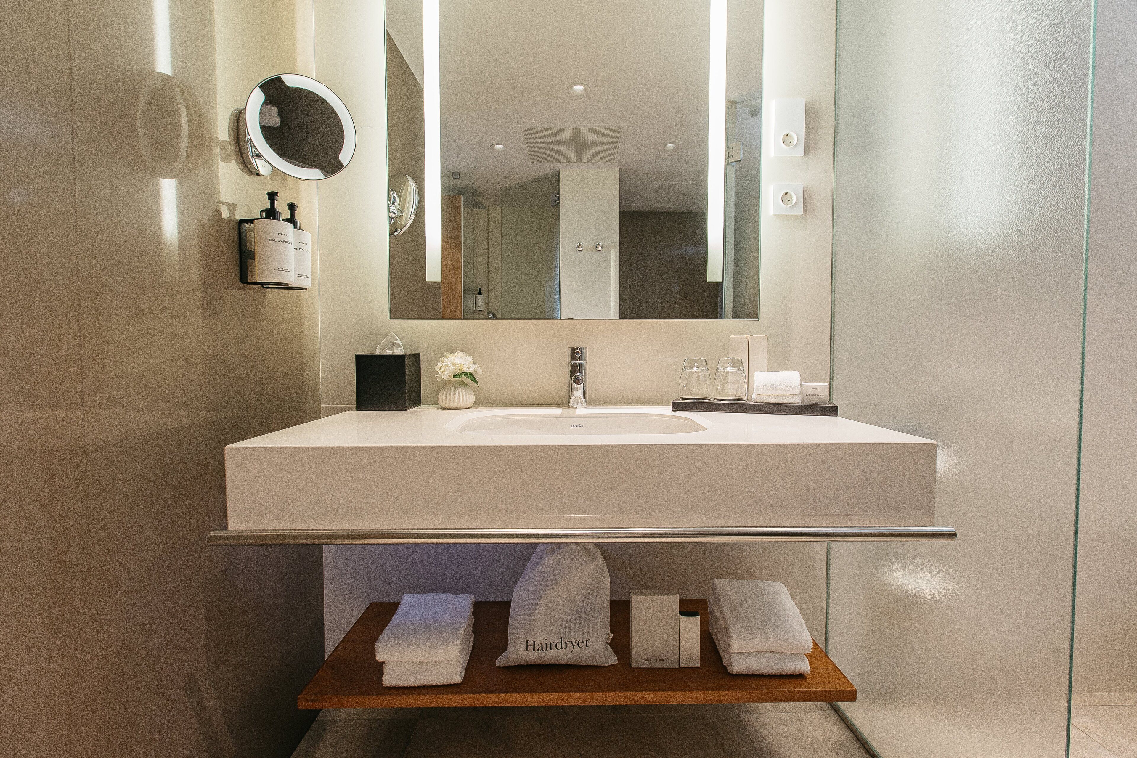 Un baño moderno con un espejo grande, lavamanos blanco y un perchero con toallas y un secador de pelo.