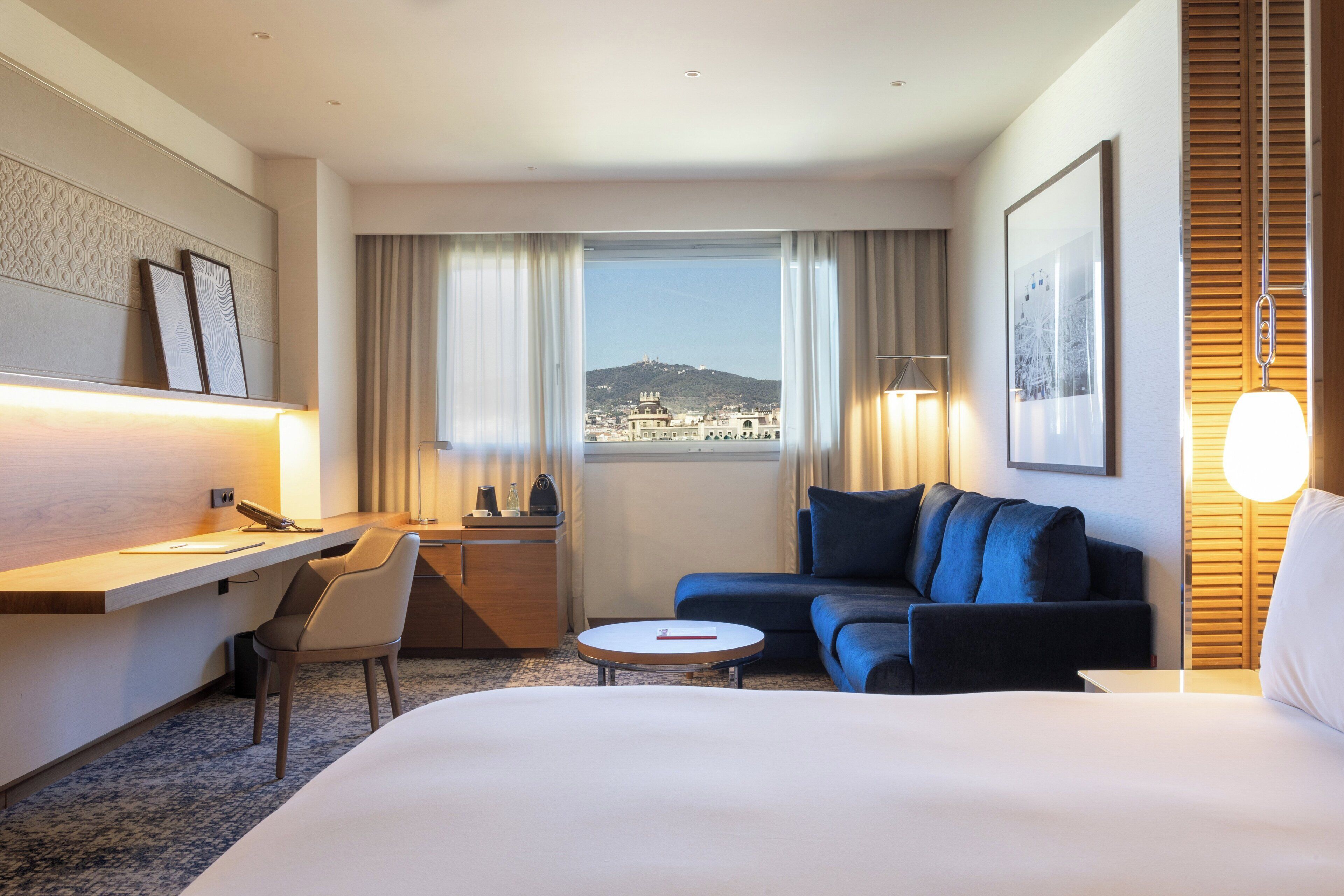 A hotel room with a bed, a desk with a chair, a sofa, a small table, and a window with a view of a cityscape.