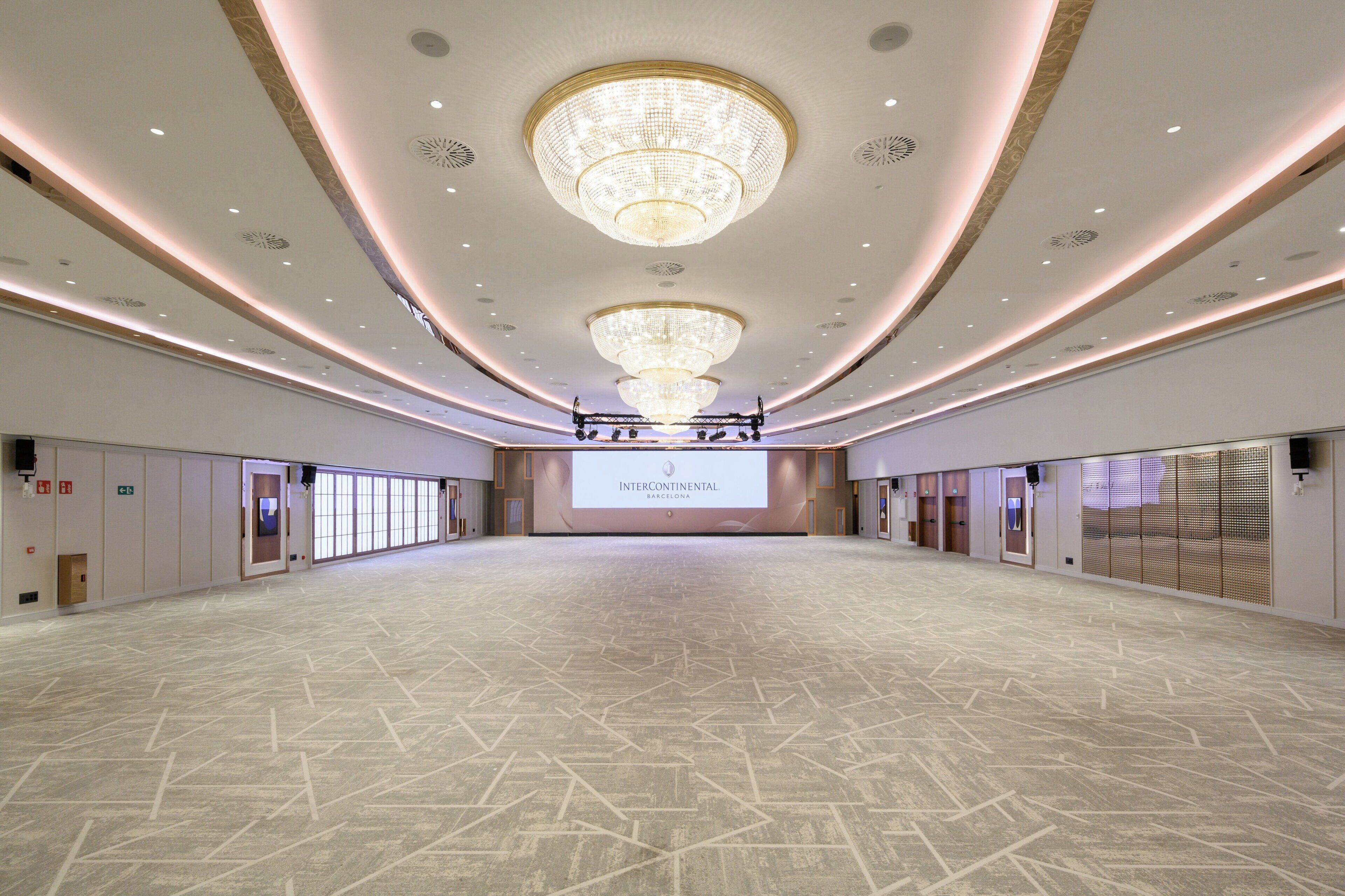 Uma sala de conferências ampla, com uma grande tela exibindo 'INTERCONTINENTAL BARCELONA', lustres e piso estampado.