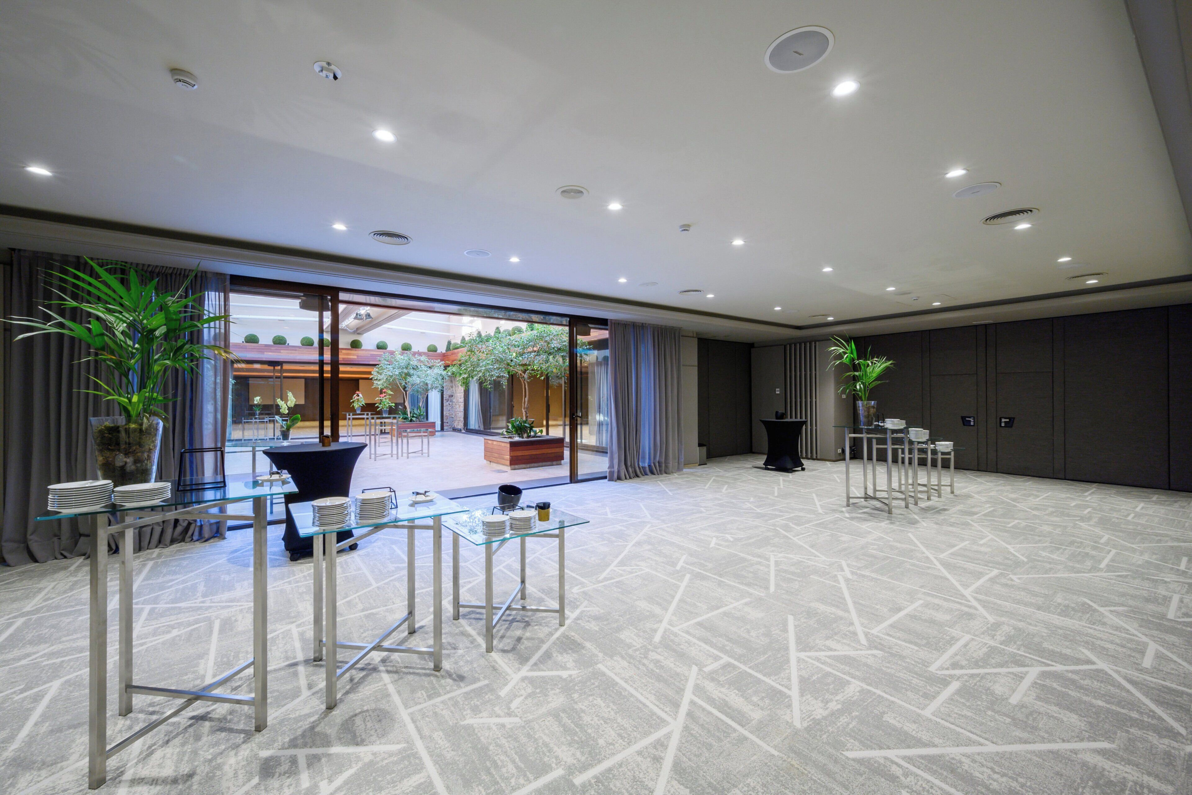 A spacious hotel lobby with a modern design, featuring a large glass partition, potted plants, and a seating area with tables and chairs.
