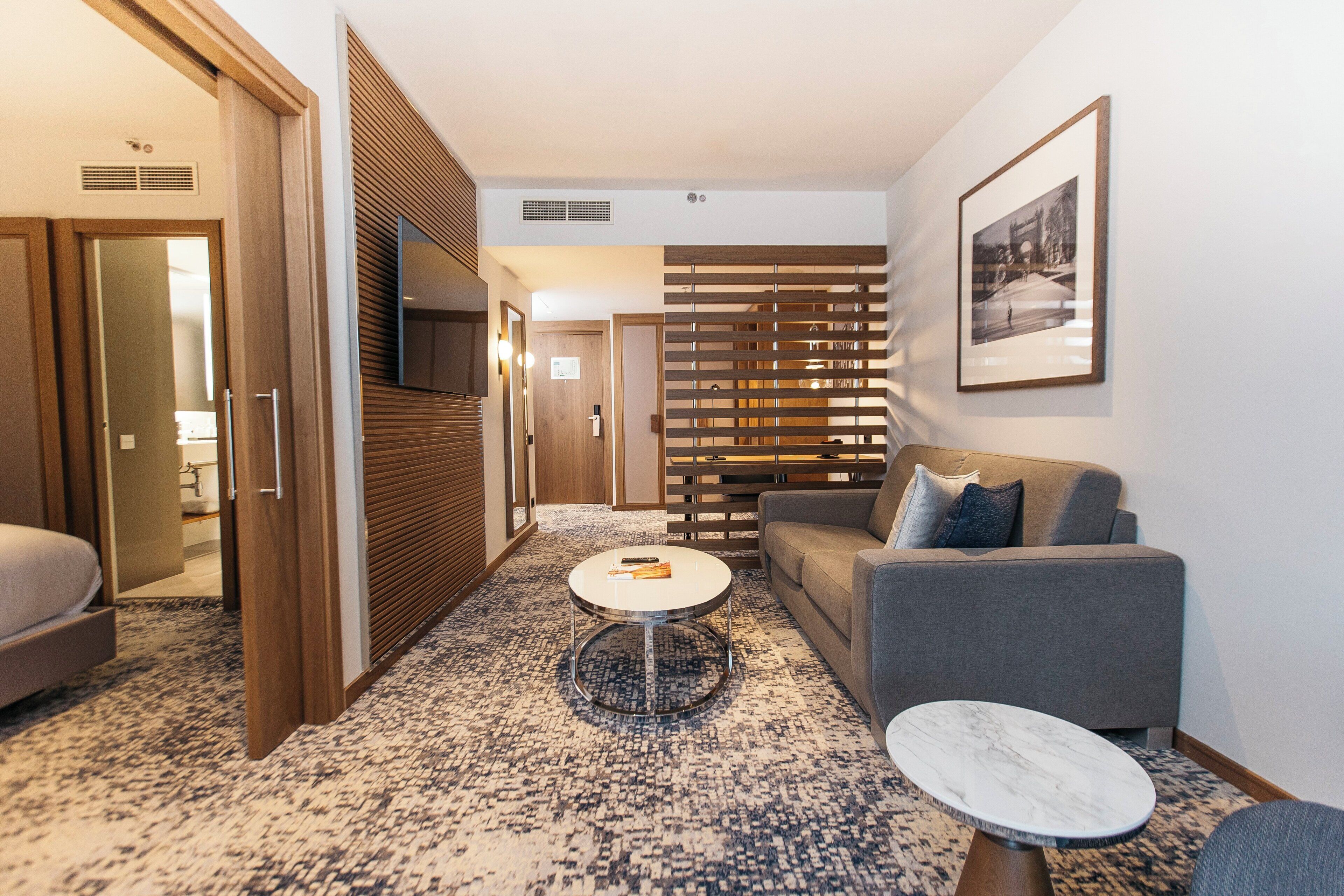 Habitación de hotel con sofá, una mesita redonda, una mesita auxiliar pequeña, alfombra estampada, panel de madera y un cuadro en la pared.