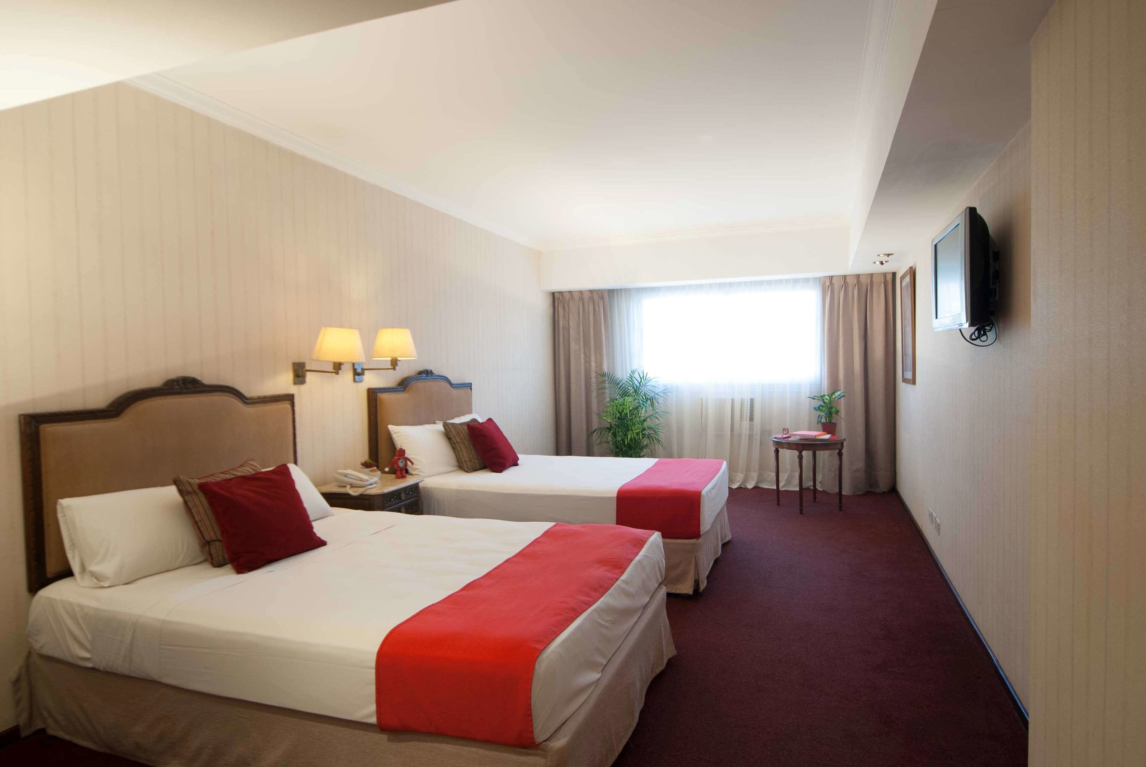 A hotel room with two beds, a television, a small table with a plant, and a window with curtains.
