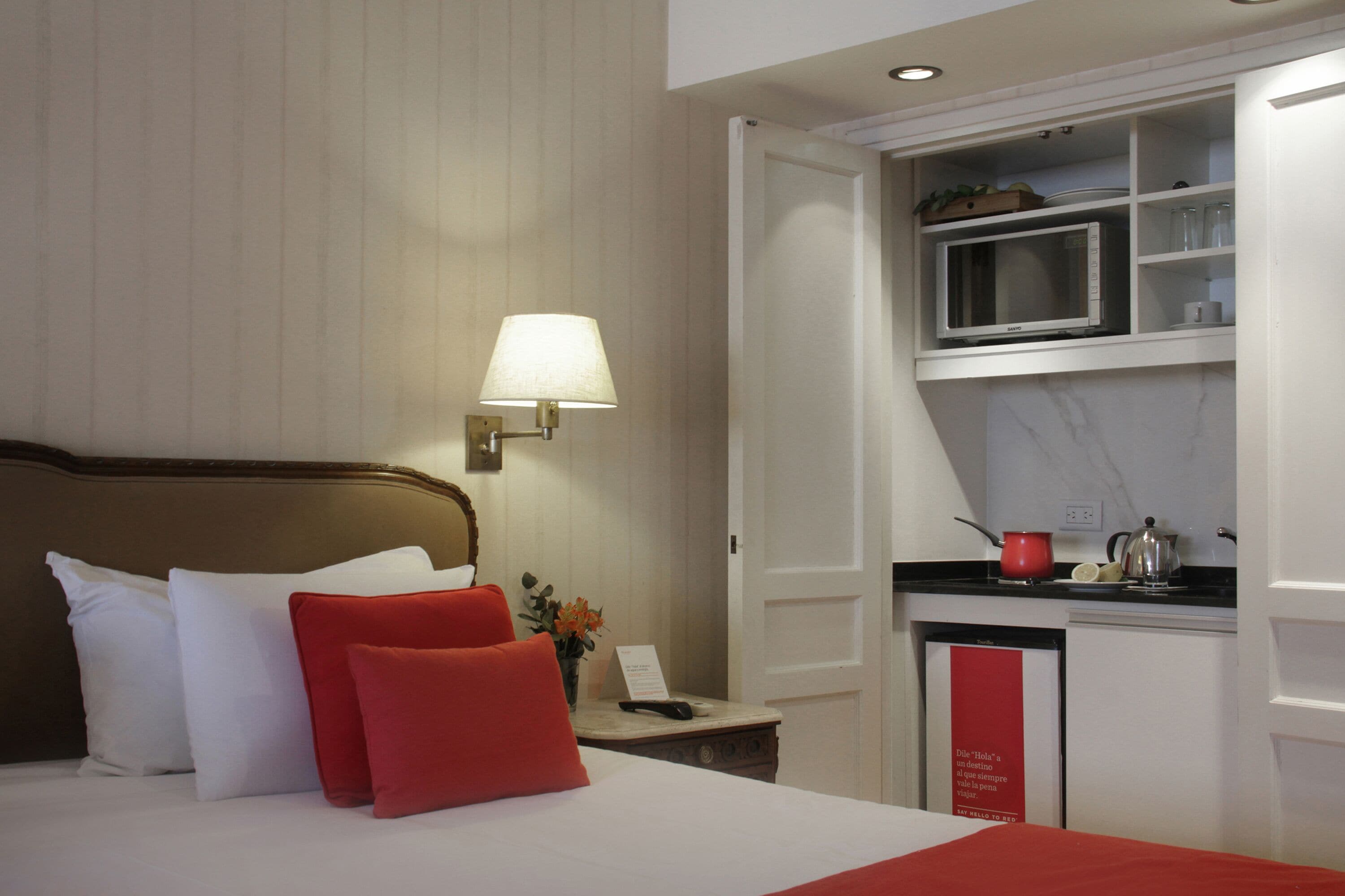 Una habitación de hotel con cama, mesita de noche, lámpara, microondas y una pequeña kitchenette.