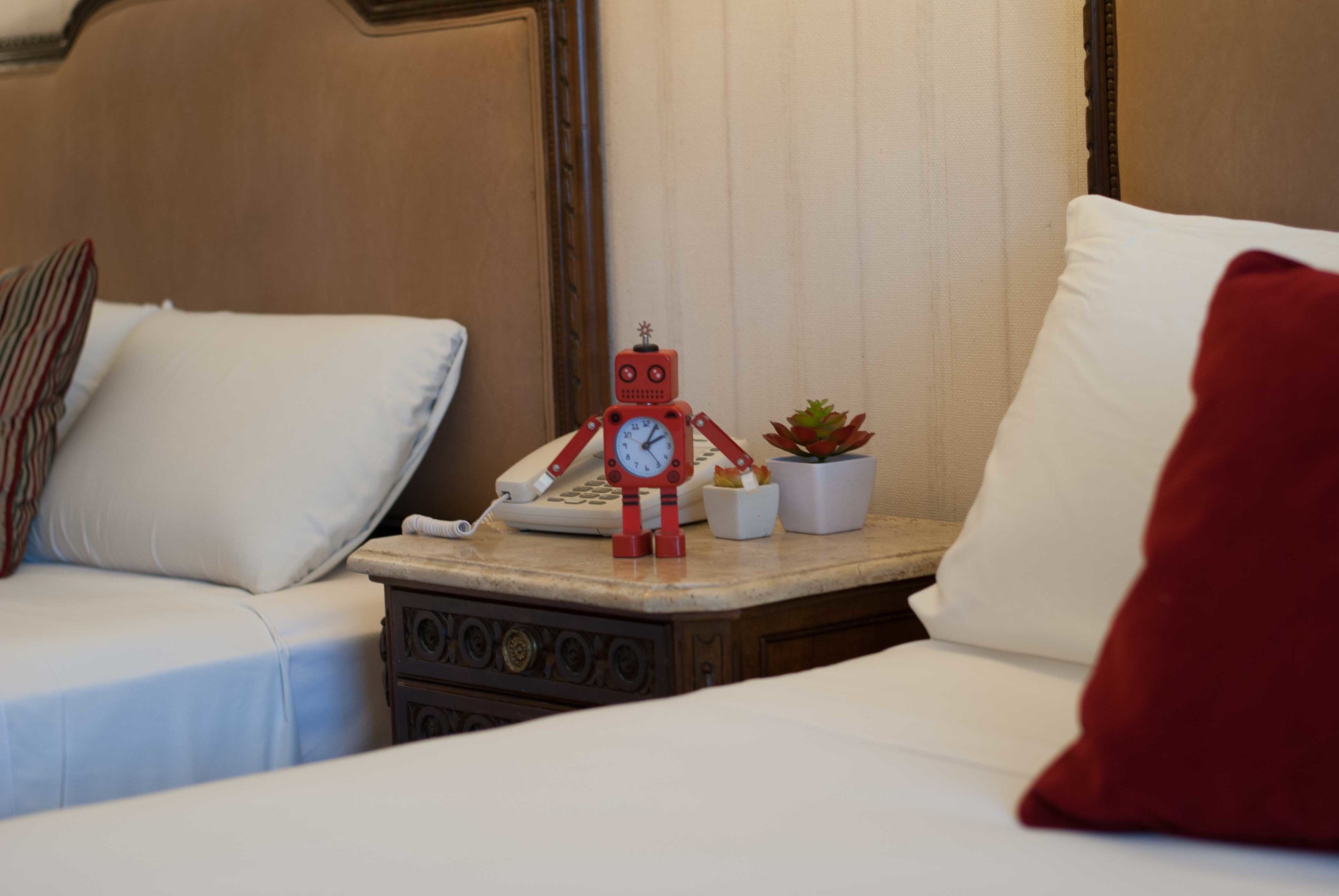 Ein Hotelzimmer mit zwei Betten, einem Nachttisch mit einem roten Roboter, einem Telefon und einer kleinen Topfpflanze.