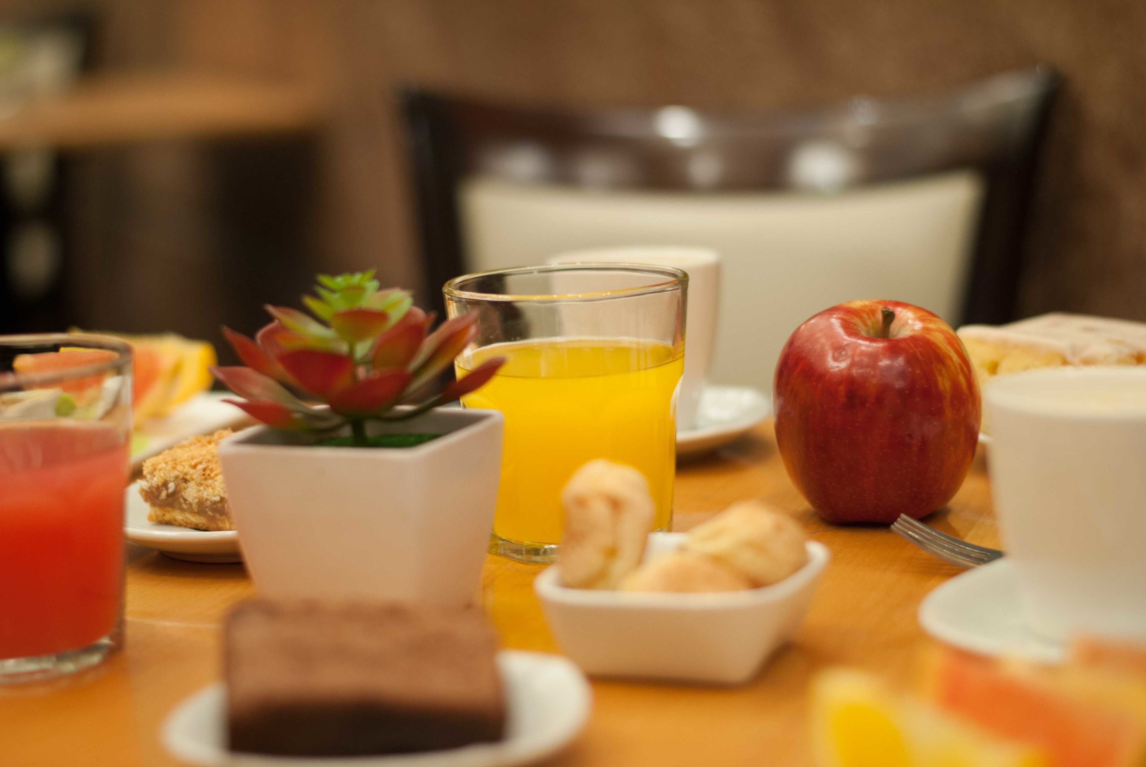 Ein Frühstückstisch mit einem Glas Orangensaft, einer Tasse Kaffee, einer Scheibe Toast, einer kleinen Schüssel Nüsse und einem roten Apfel.