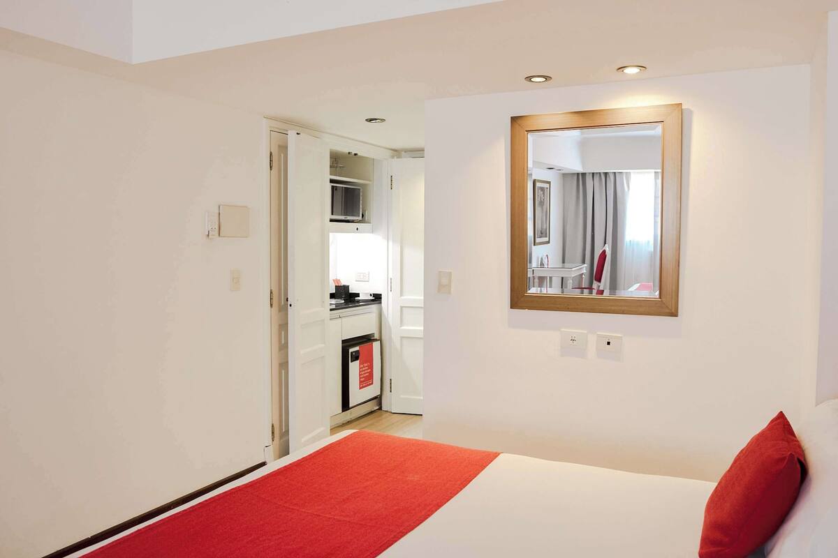 A hotel room with a bed, a red throw, a mirror, and a view into the kitchen.