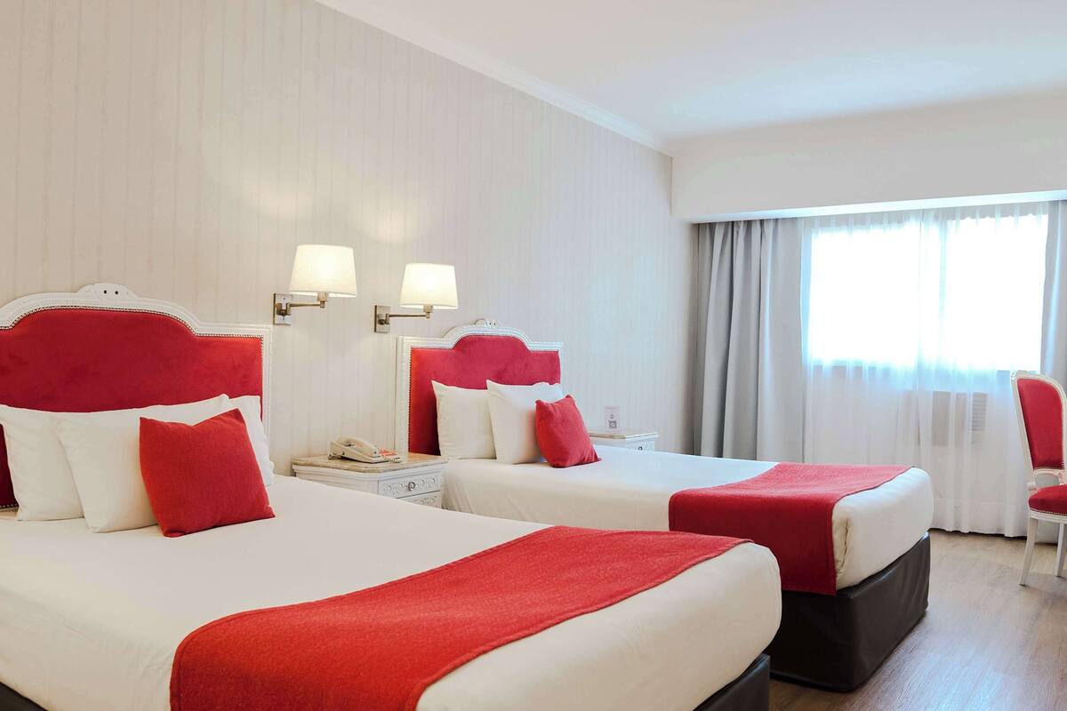 A hotel room with two beds, each with a red headboard and white bedding. There are two bedside lamps, a window with curtains, and a chair.