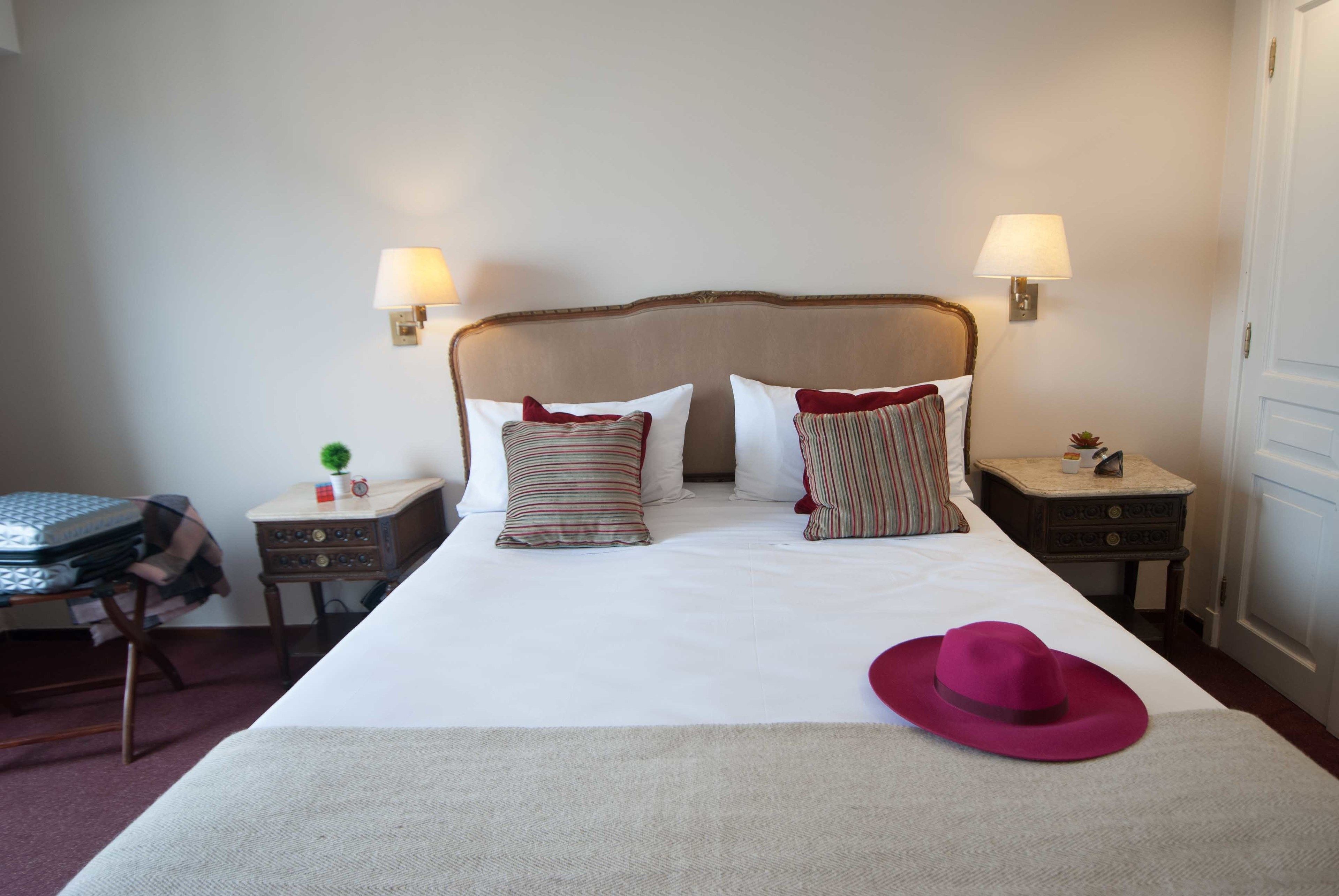A neatly made bed with a striped pillow, a red hat on the bed, and two bedside tables with lamps.