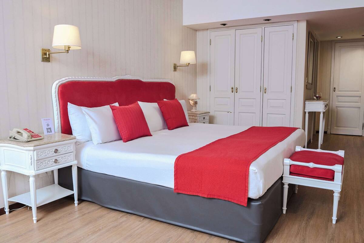 A large bed with a red headboard, white bedding, and a red throw blanket. There are two bedside tables with lamps, a small bedside table with a lamp, and a white nightstand with a phone.