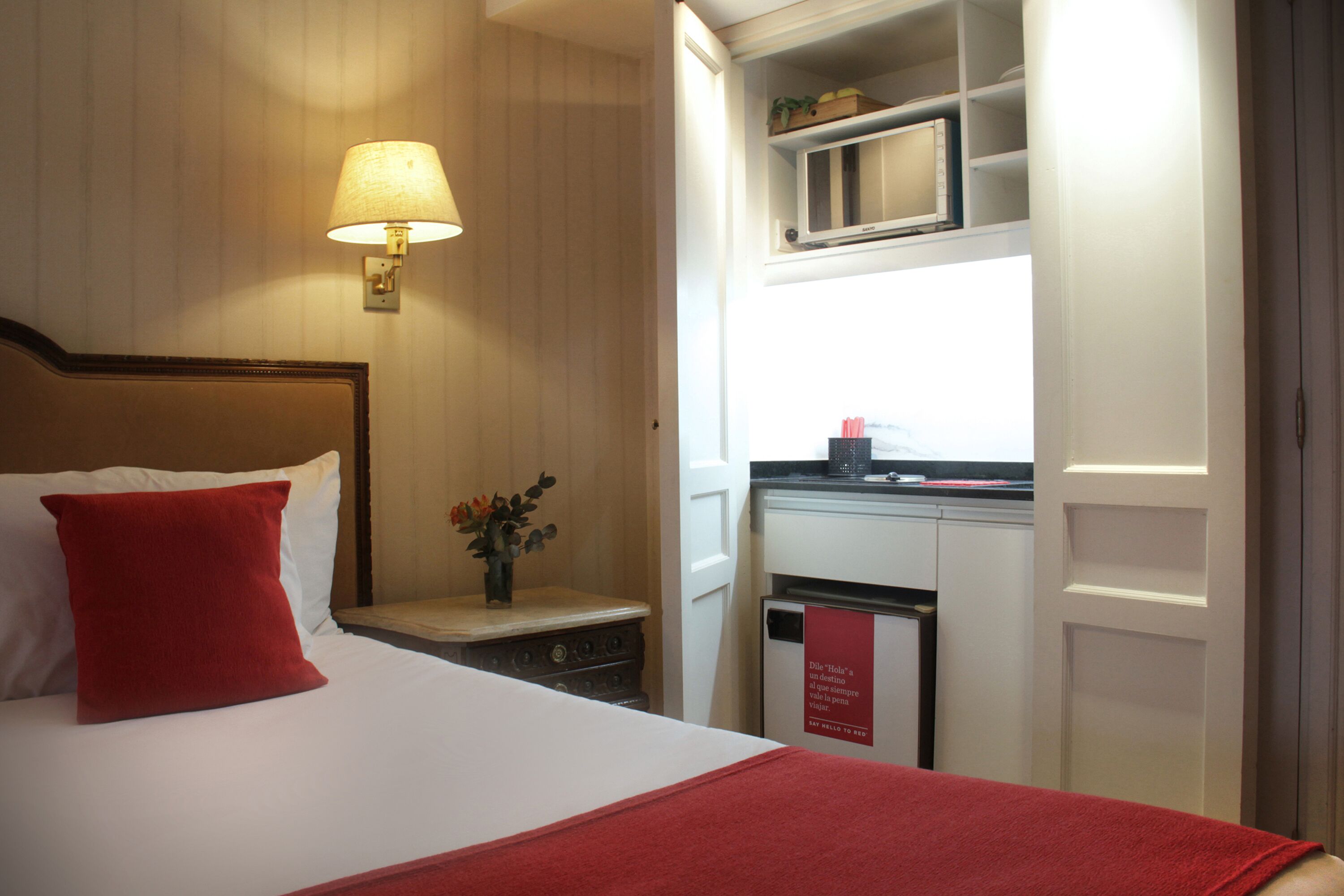 A hotel room with a bed, a nightstand, a lamp, a microwave, and a small kitchen area.