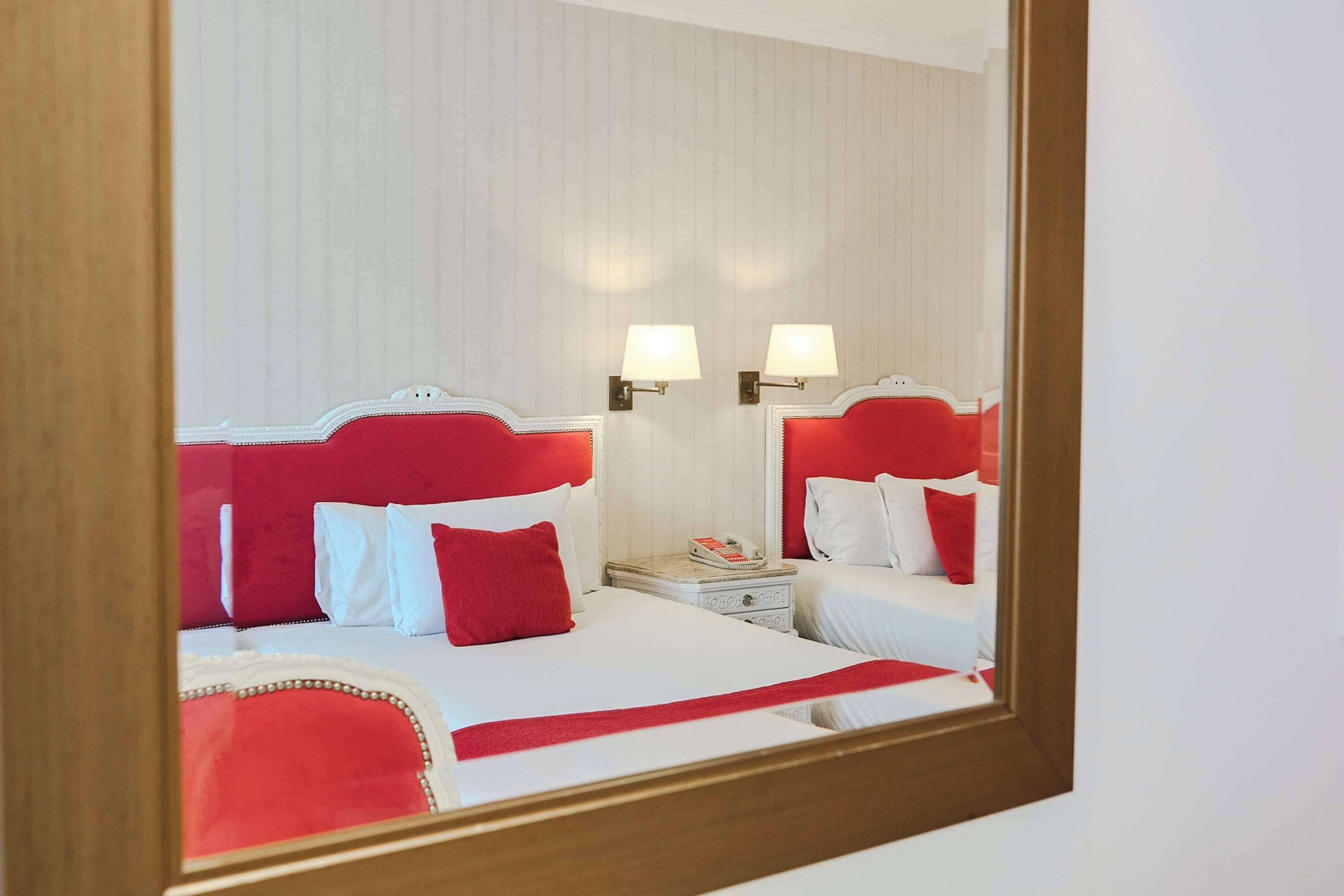 A hotel room with two beds, each with a red headboard, white bedding, and a red throw blanket. There are two bedside tables with lamps and a telephone.