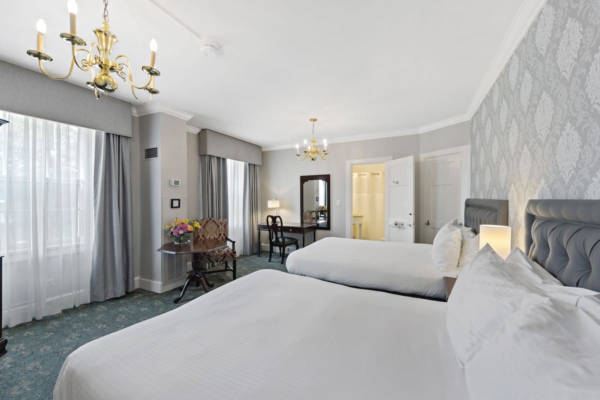 A hotel room with two beds, a desk with a chair, a chandelier, and a large mirror.