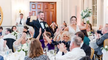 A formal event with guests seated at round tables, clapping, and a couple in the foreground.
