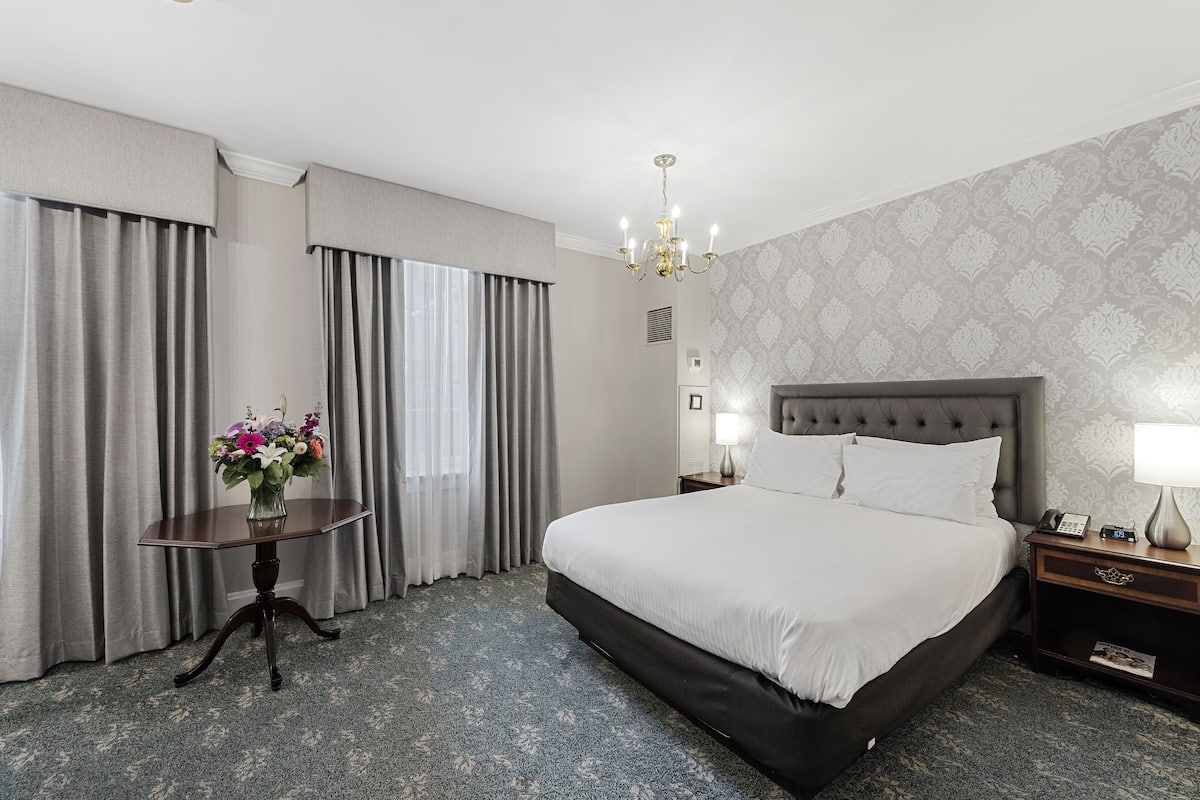 A hotel room with a large bed, a bedside table with a lamp, a nightstand, and a vase of flowers.