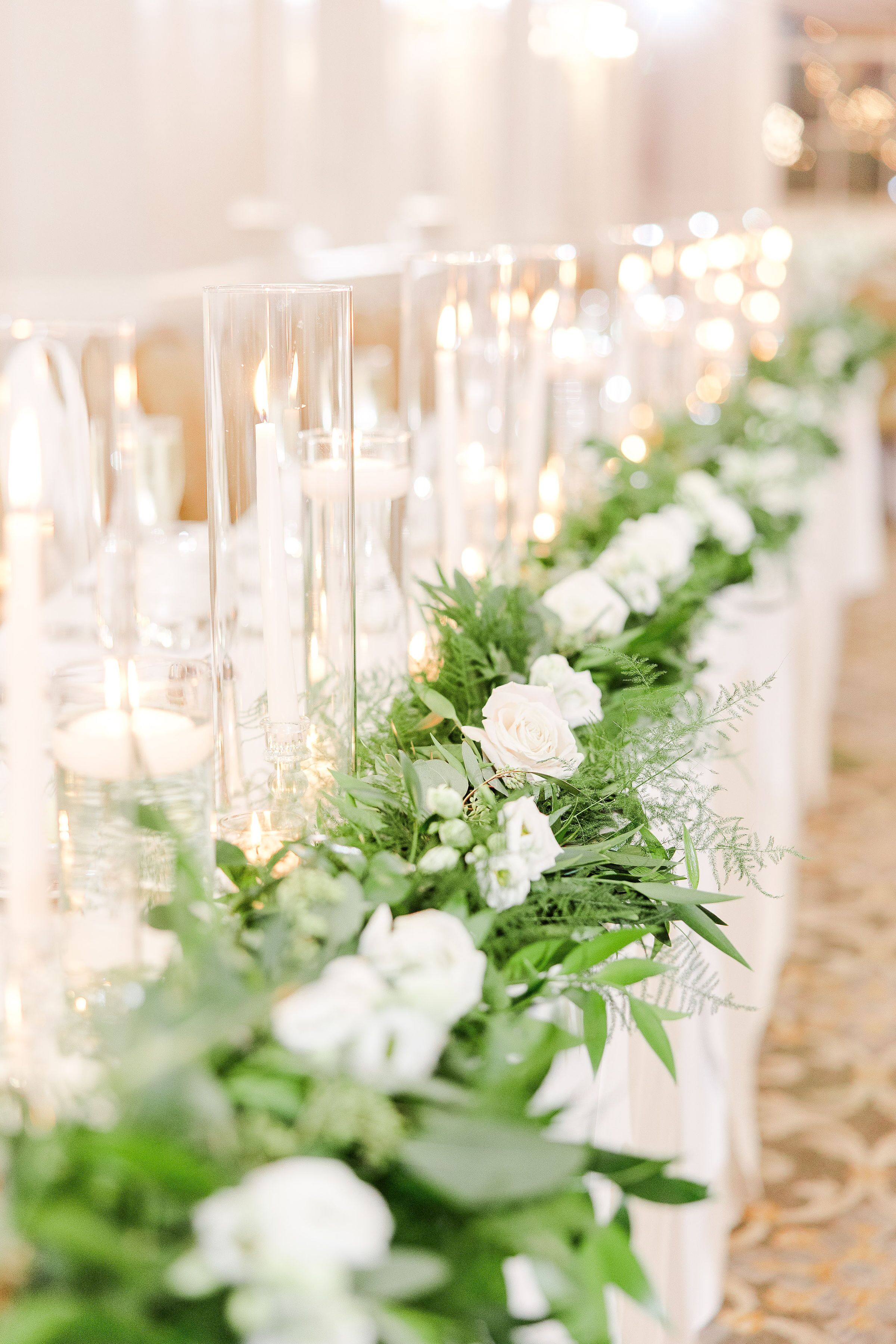 A long table with clear glass candle holders, white flowers, and greenery, likely set up for a formal event.