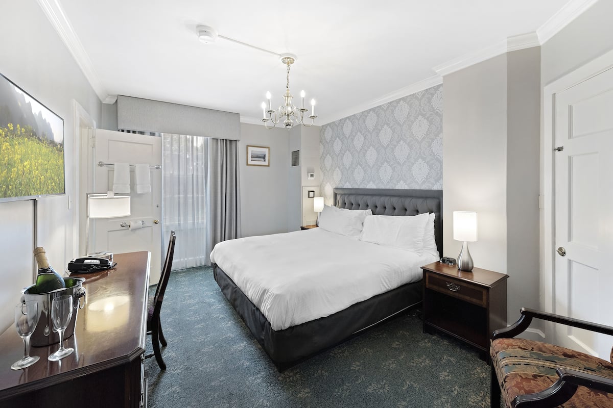 A hotel room with a large bed, a bedside table, a chair, a desk with a bottle of champagne, and a lamp.