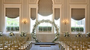 An indoor venue with a central aisle, flanked by rows of chairs, and a decorative floral arch at the front. - Hawthorne Hotel (Salem)