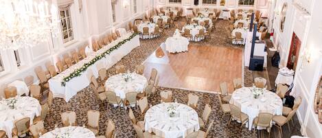 A spacious banquet hall with multiple round tables, each set with white tablecloths and gold chairs, arranged in a symmetrical layout.