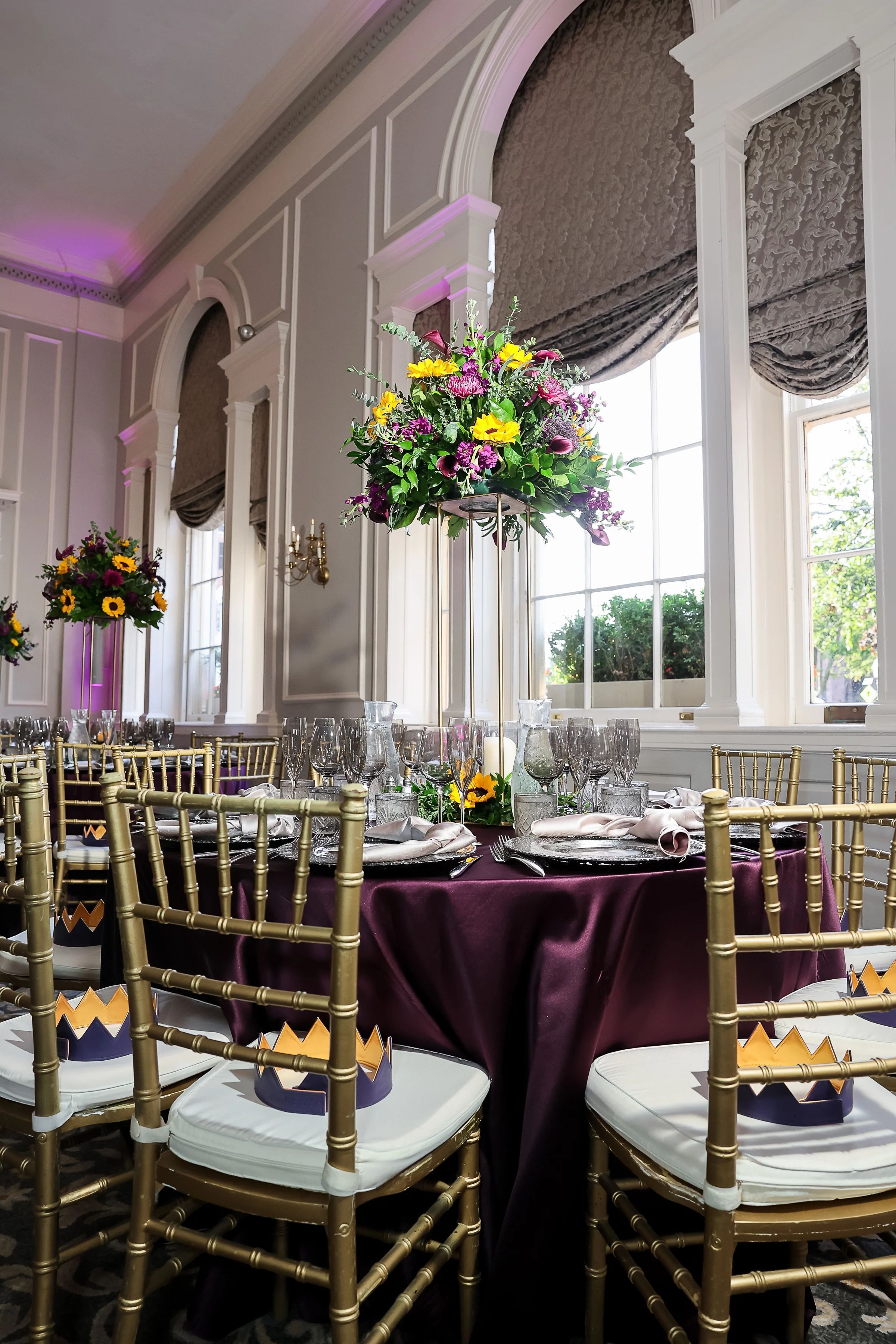 Un salón de banquetes con sillas doradas, manteles morados y centros de mesa florales.