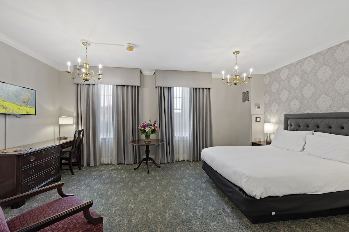 A hotel room with a large bed, a desk, a chair, a small table with a vase of flowers, and a painting on the wall.