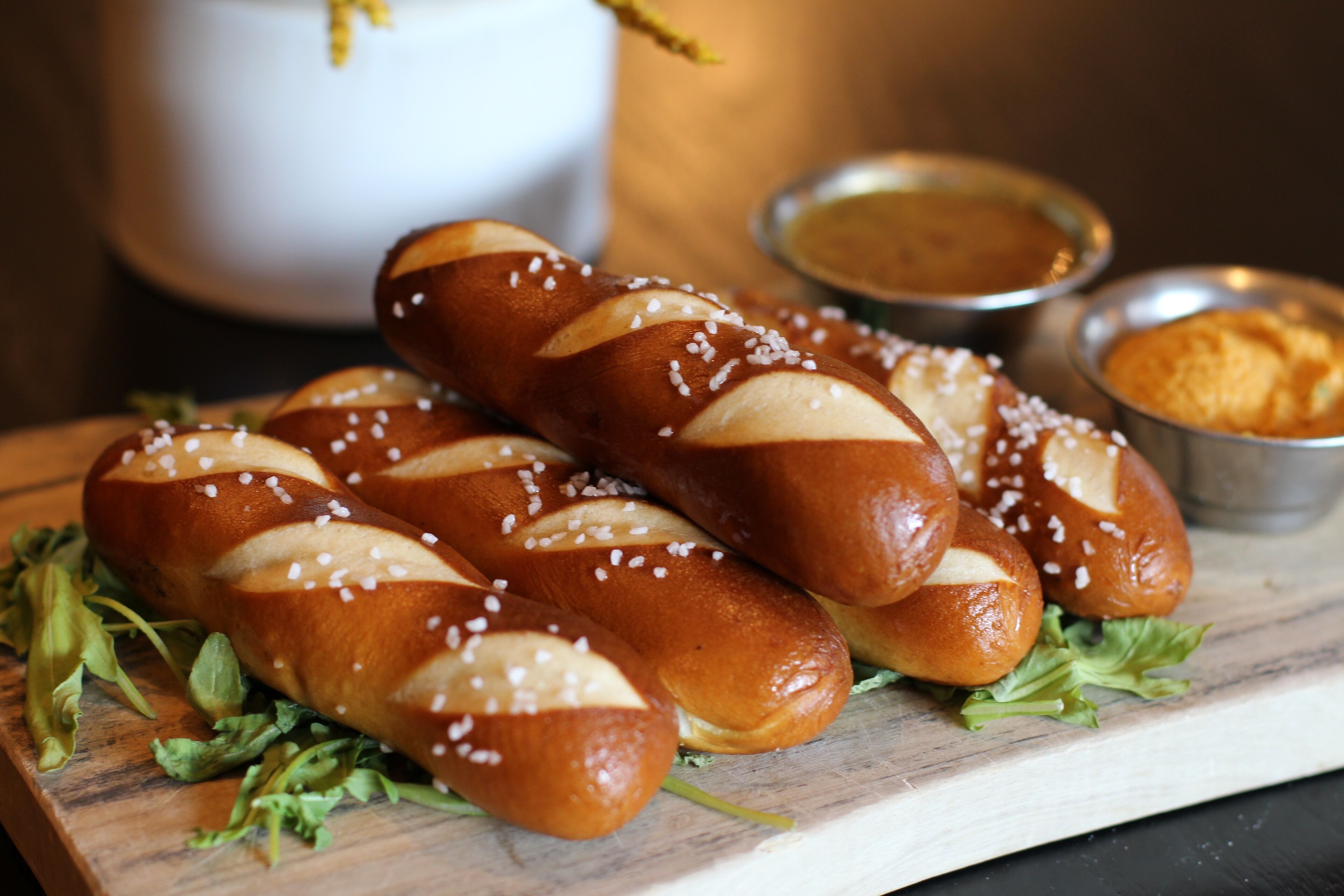 Una tabla de madera con varios panecillos tipo pretzel, algunos con sal, y pequeños recipientes metálicos con salsas para mojar.