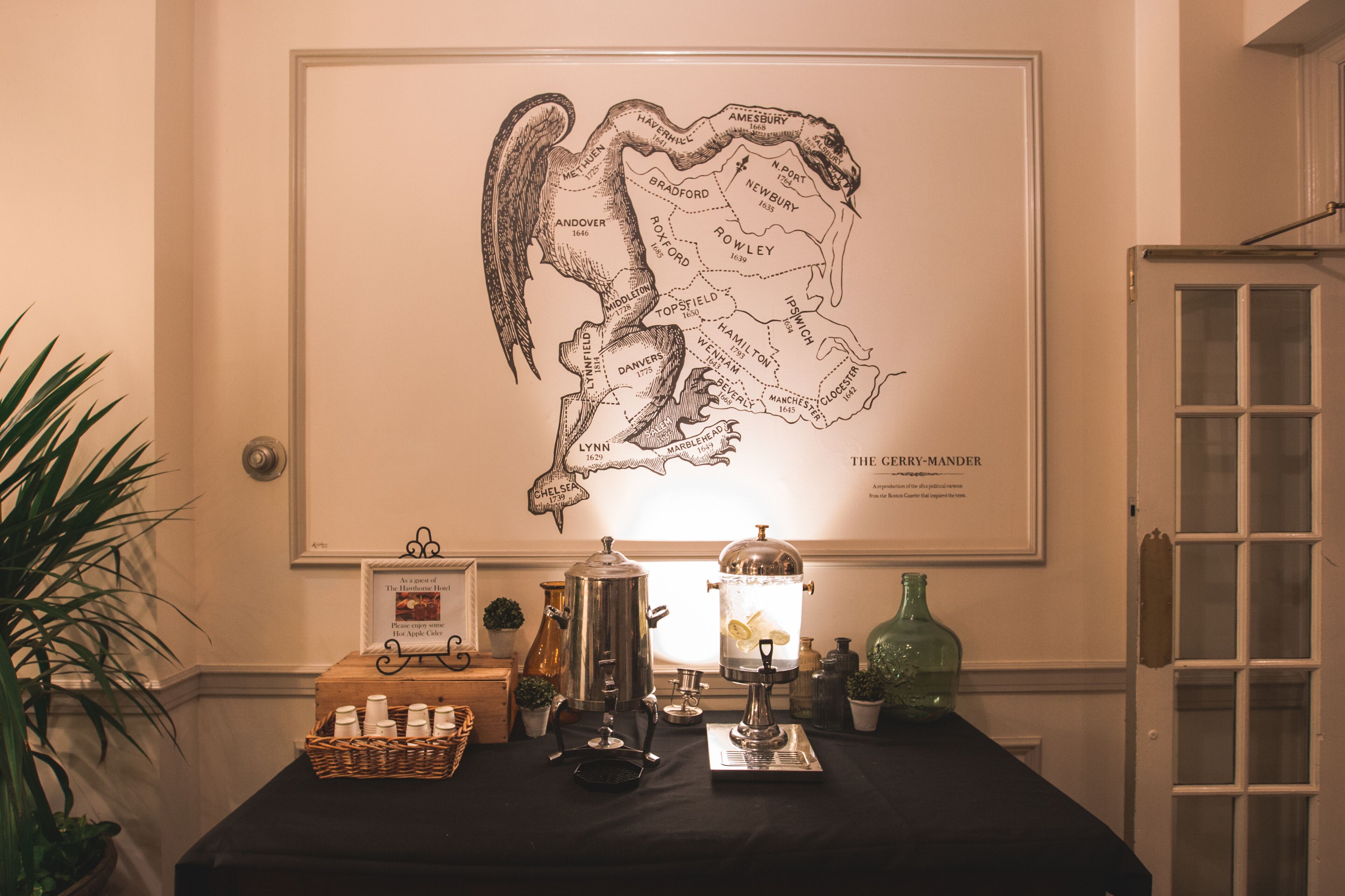 Un mapa enmarcado del Reino Unido con un borde decorativo, una mesa con una tetera, hervidor y botellas, y una planta en maceta a la izquierda.