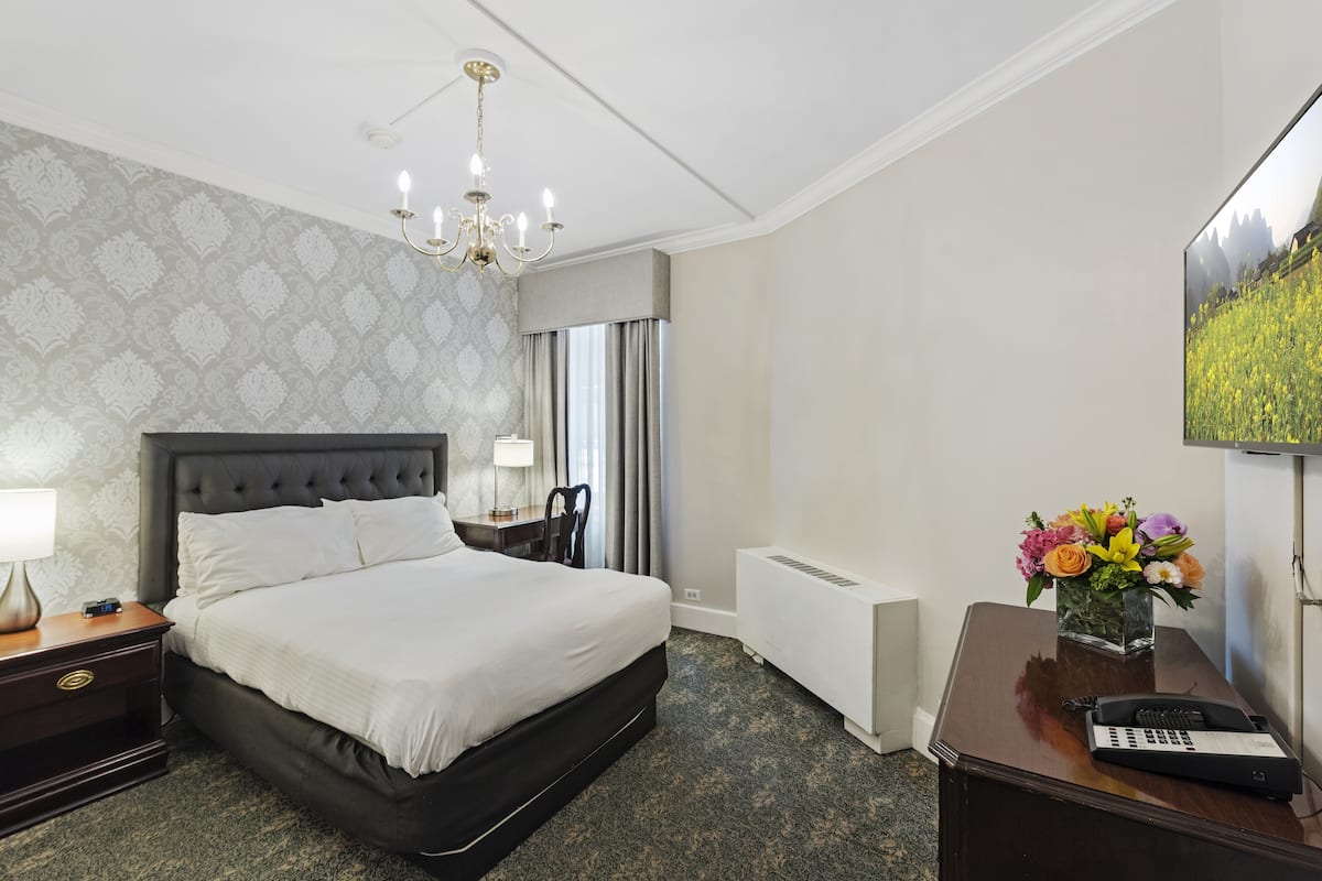 A hotel room with a large bed, a desk with a telephone, a chair, a nightstand with a lamp, a radiator, and a wall-mounted TV.