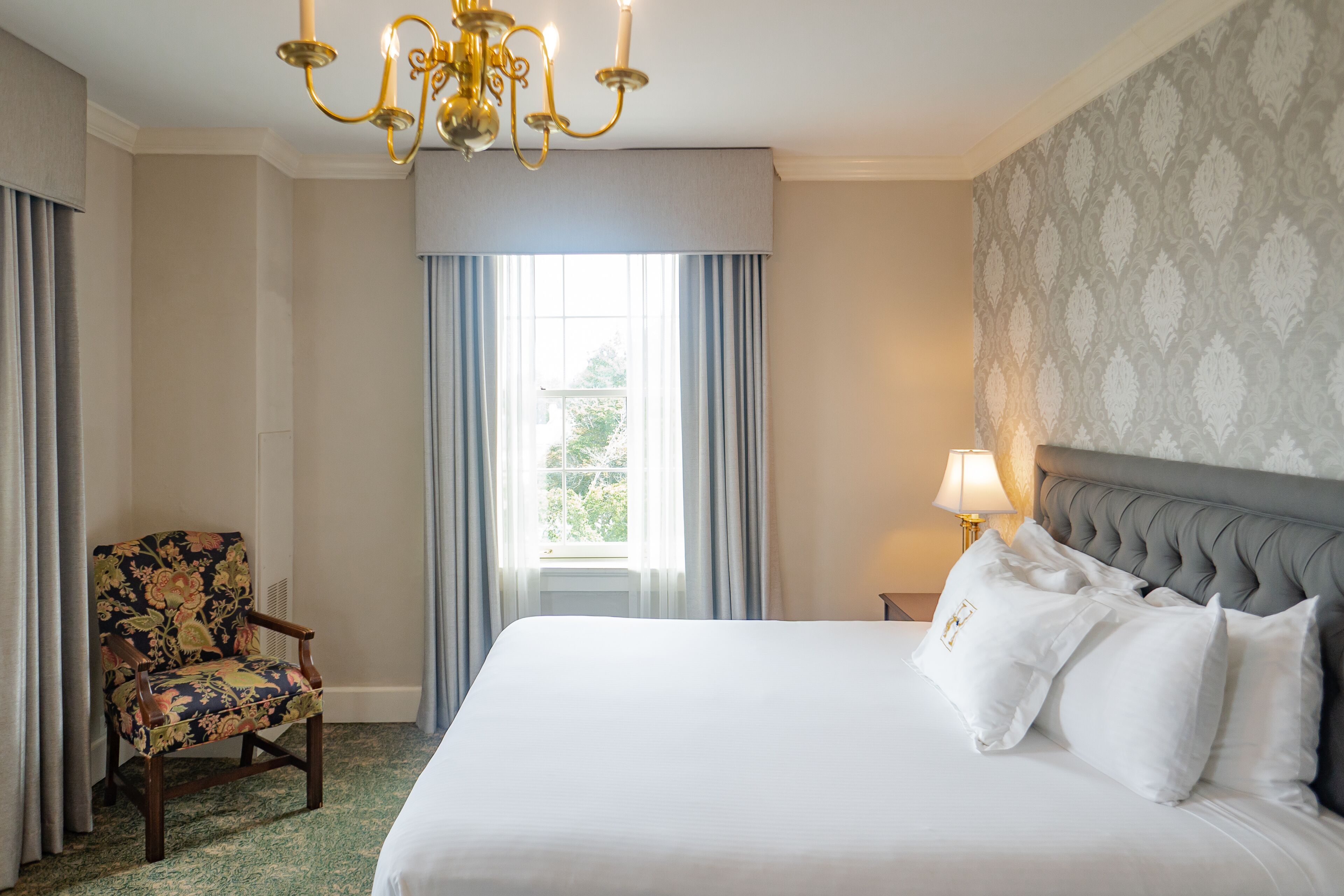 Une chambre d’hôtel avec un grand lit, une chaise, un lustre et une fenêtre avec des rideaux.