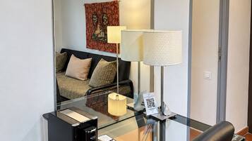 A hotel room with a glass desk, a coffee maker, a telephone, a lamp, and a bed with a patterned headboard.