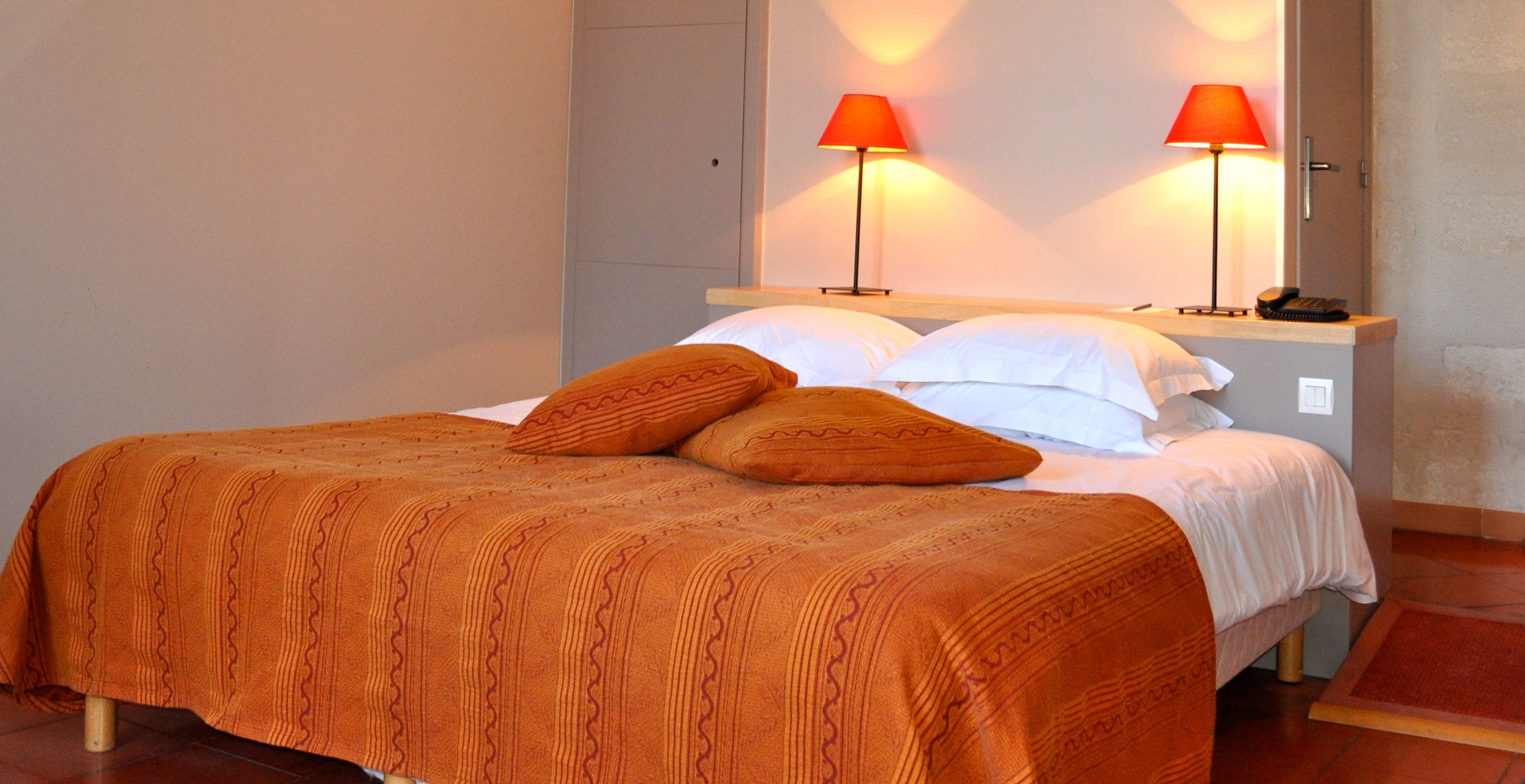 Ein Bett mit einem gemusterten, orangefarbenen Überwurf, zwei Kissen und zwei rote Lampen auf einem Nachttisch.