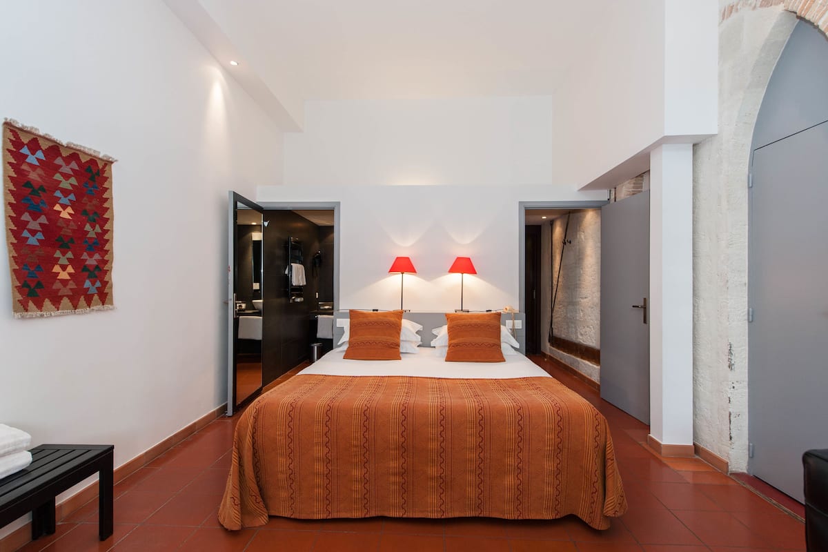 Una habitación de hotel con una cama grande, dos lámparas rojas, un banco y una alfombra colorida.