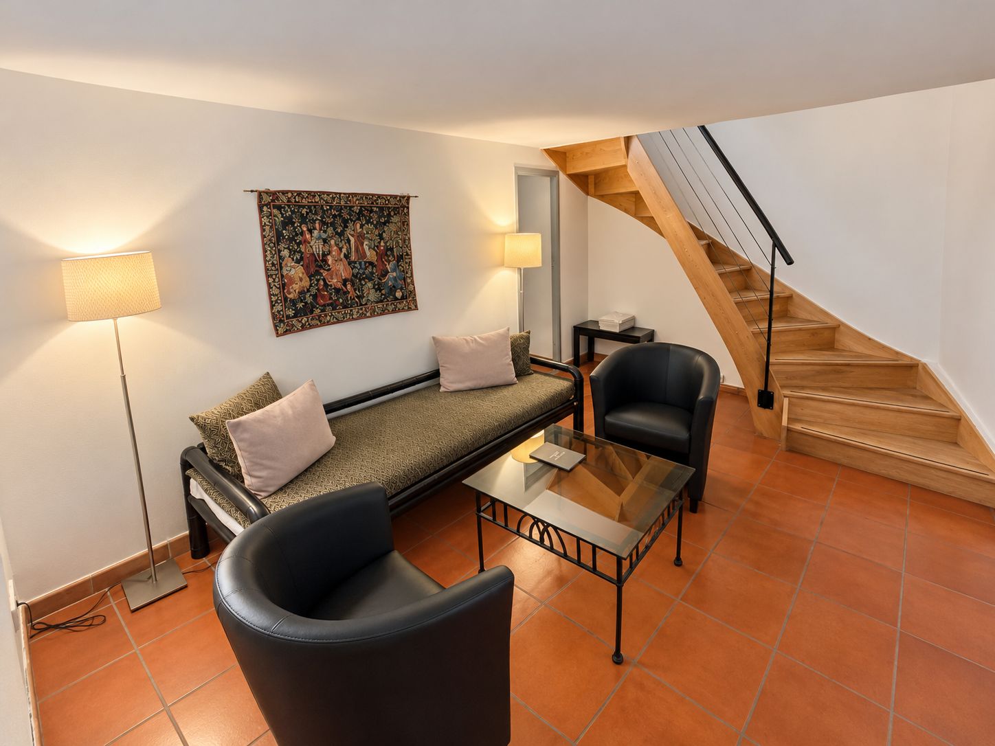 Een moderne woonkamer met een bank, een glazen salontafel, een zwarte fauteuil, een trap en een wandtapijt.