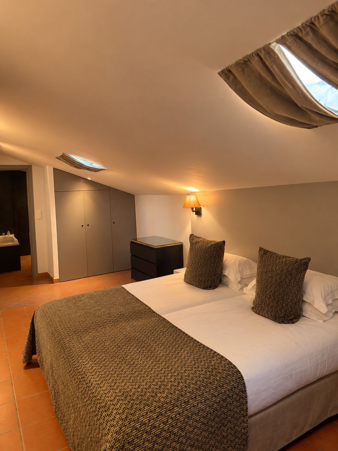 Een slaapkamer met een groot bed, twee kussens, een nachtkastje, een lamp, een kledingkast en een badkamer die door een open deur zichtbaar is.
