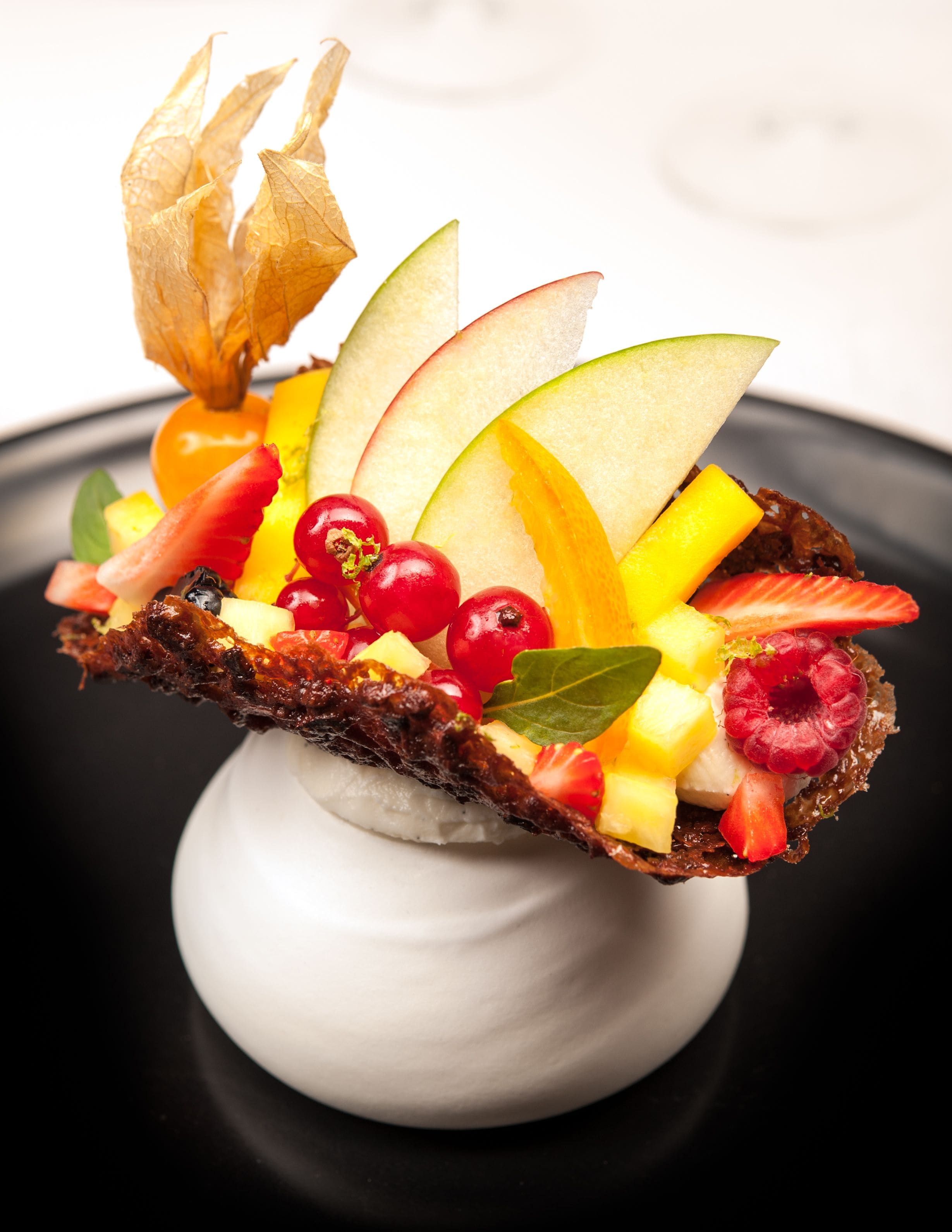 A dessert with various fruits and a piece of burnt toast on a white pedestal.