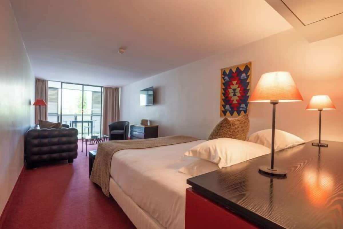 Habitación de hotel con cama, dos lámparas de noche, escritorio, sillón, sofá y ventana con cortinas.