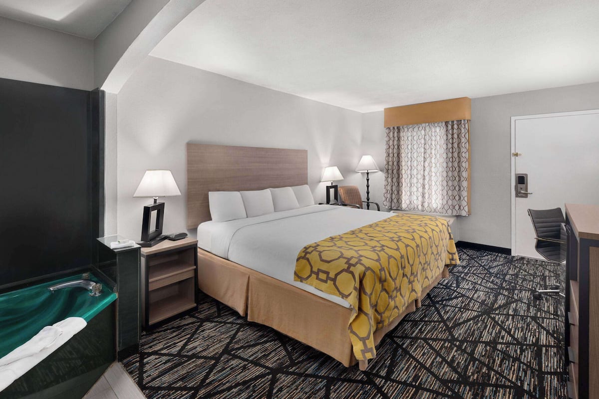 A hotel room with a bed, two bedside lamps, a desk, a chair, a TV, and a bathroom with a bathtub.