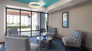 Un hall d’entrée moderne avec un tapis bleu, deux fauteuils gris, une table basse ronde surmontée d’un vase bleu, et une grande fenêtre offrant une vue sur des immeubles et un parking.