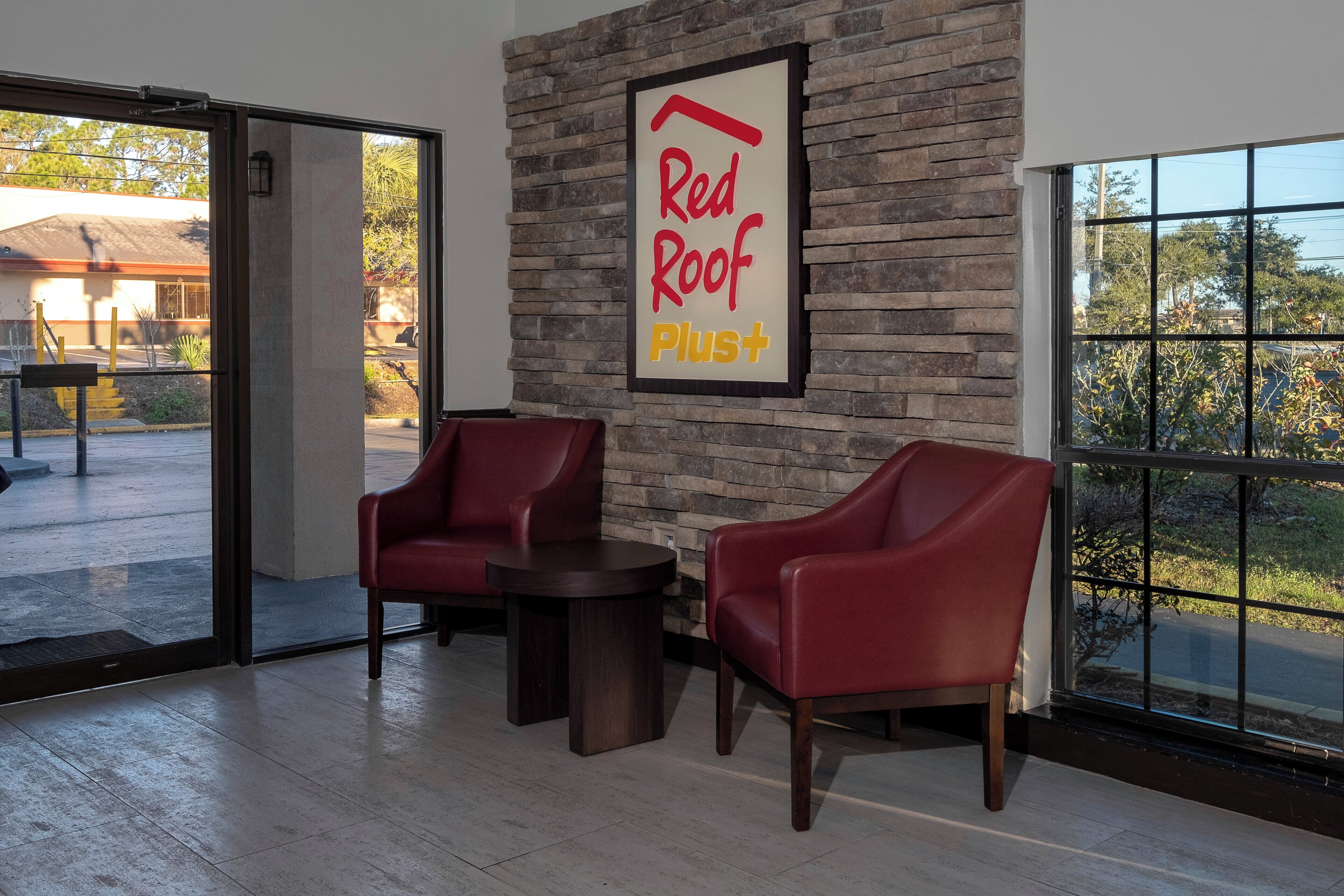 Une pièce avec un mur en pierre, une enseigne « Red Roof Plus », deux chaises rouges, une petite table et une grande fenêtre donnant sur des arbres.