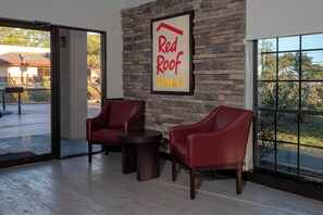 A room with a stone wall, a Red Roof Plus sign, two red chairs, a small table, and a large window with a view of trees. - Red Roof Inn PLUS+ St Augustine (St. Augustine)